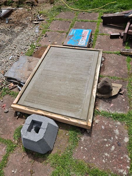 A concrete slab in a wooden frame sits on paving stones, a gray block and a saw nearby. Construction setting.