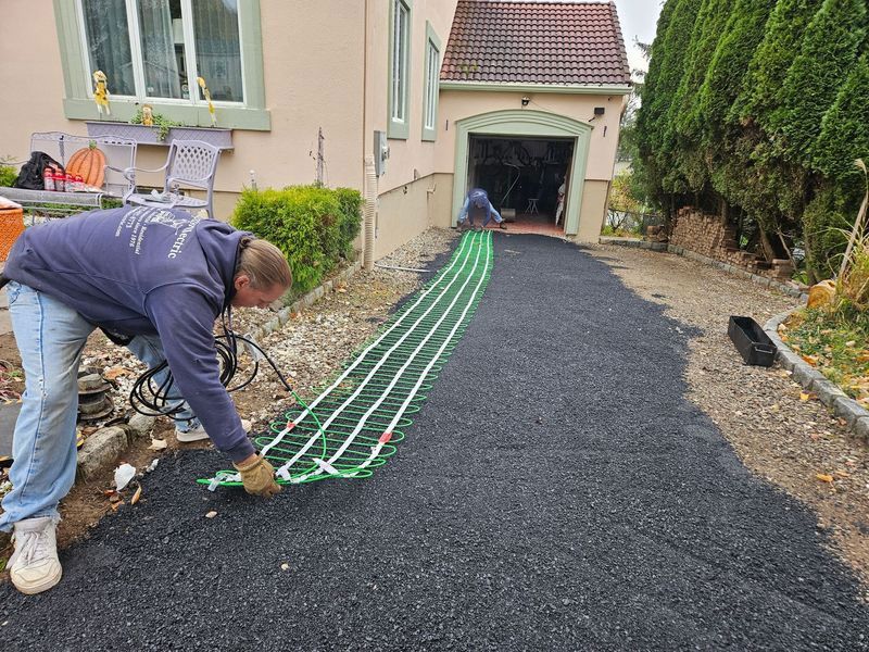 Two men installing a heated driveway, with a green cable system on dark asphalt in front of a garage.