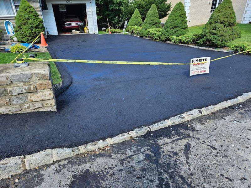Freshly paved black asphalt driveway, bordered by stone and grass, with a garage in the background.