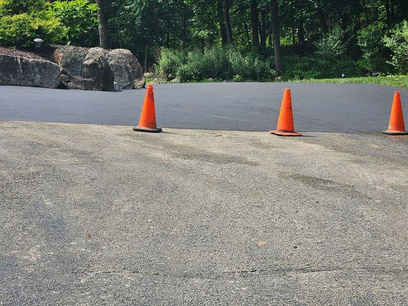 A freshly paved asphalt driveway with orange cones marking the new section, contrasted with the older, weathered surface.