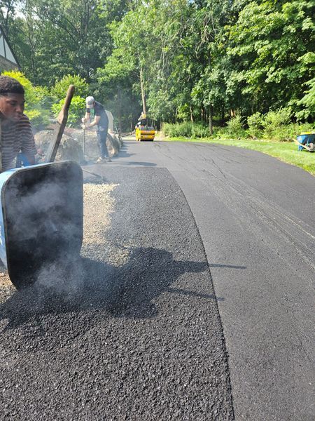 Road workers paving a curved driveway, using a roller, shovels, and a wheelbarrow. Sunny day with trees in the background.