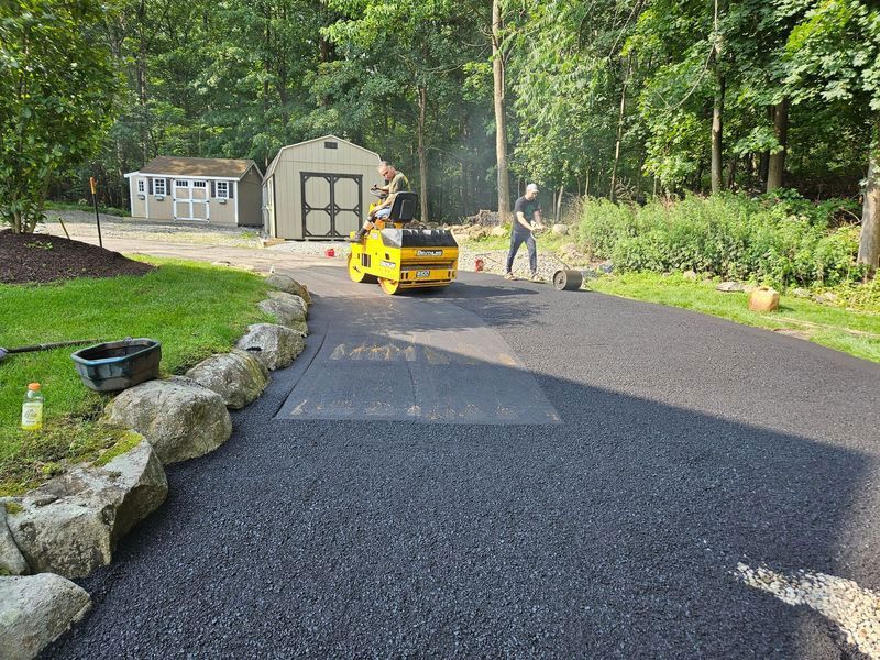New asphalt driveway being paved with a yellow roller machine and a worker in a wooded residential setting.