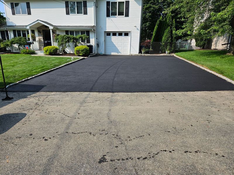 Newly paved black asphalt driveway in front of a two-story white house with a lawn.