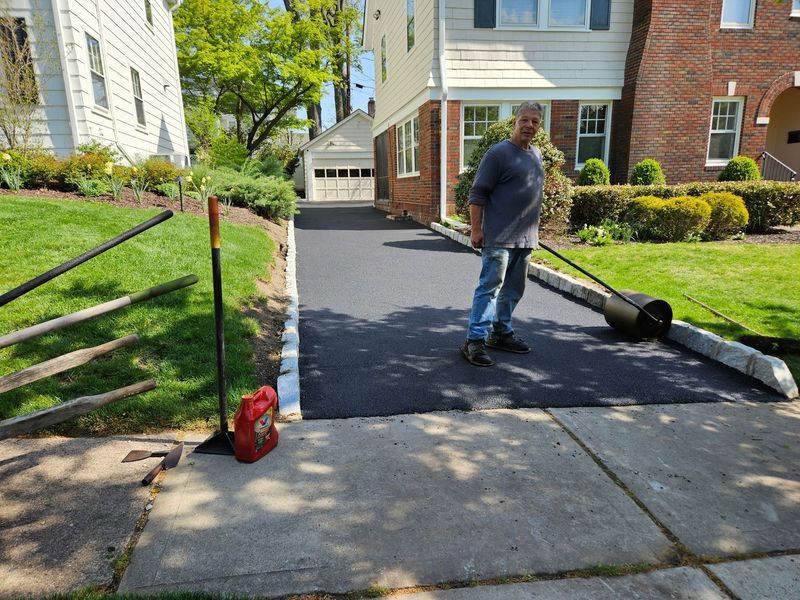 Man standing on a newly paved driveway, using a roller. House and garage in the background, sunny day.