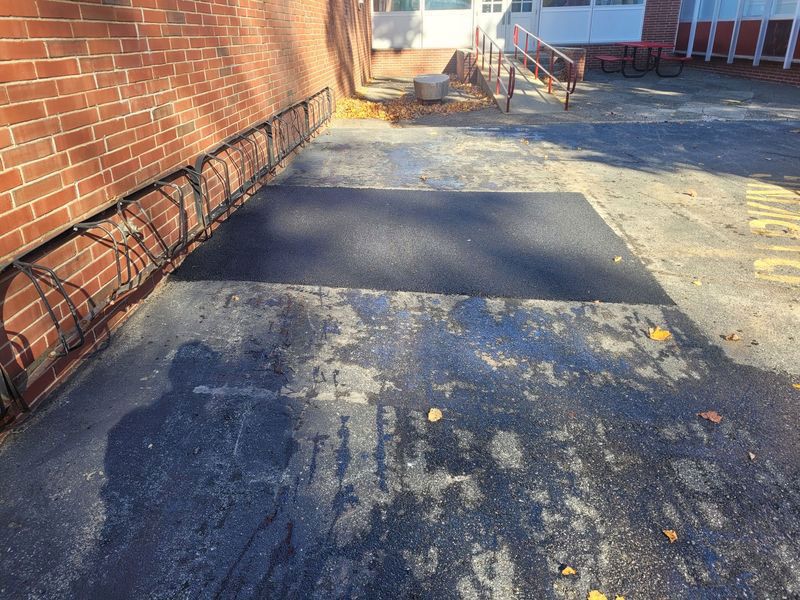 A rectangular patch of fresh asphalt repairs the pavement next to a red brick wall, near a building entrance with a ramp.