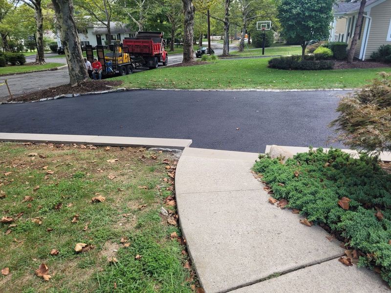 Newly paved black asphalt driveway in front of a house, with a concrete walkway. A paving truck is visible in the background.