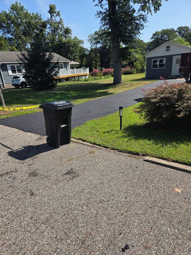 Blue recycling bin on a newly paved driveway in front of a garage. A trailer with equipment is in the background.