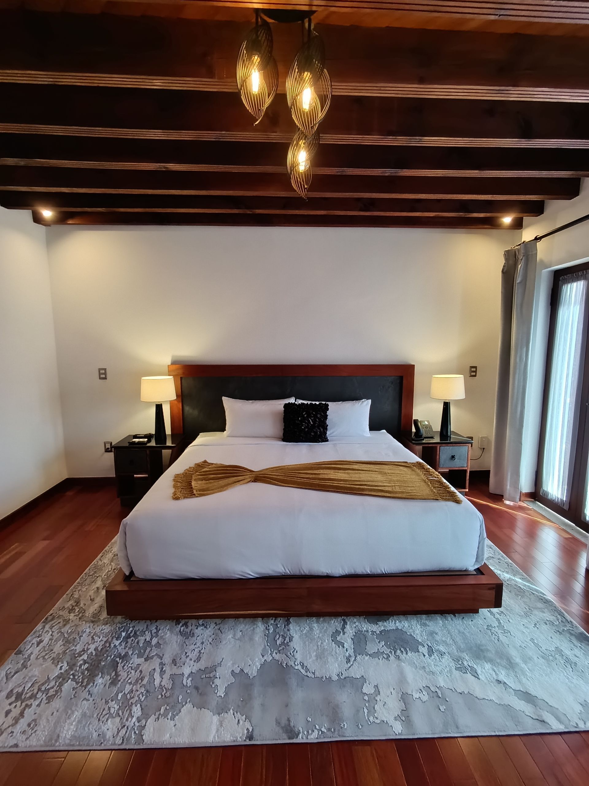 Dormitorio con cama de madera, ropa de cama blanca y una manta dorada. Lámparas colgantes, vigas de madera y alfombra.
