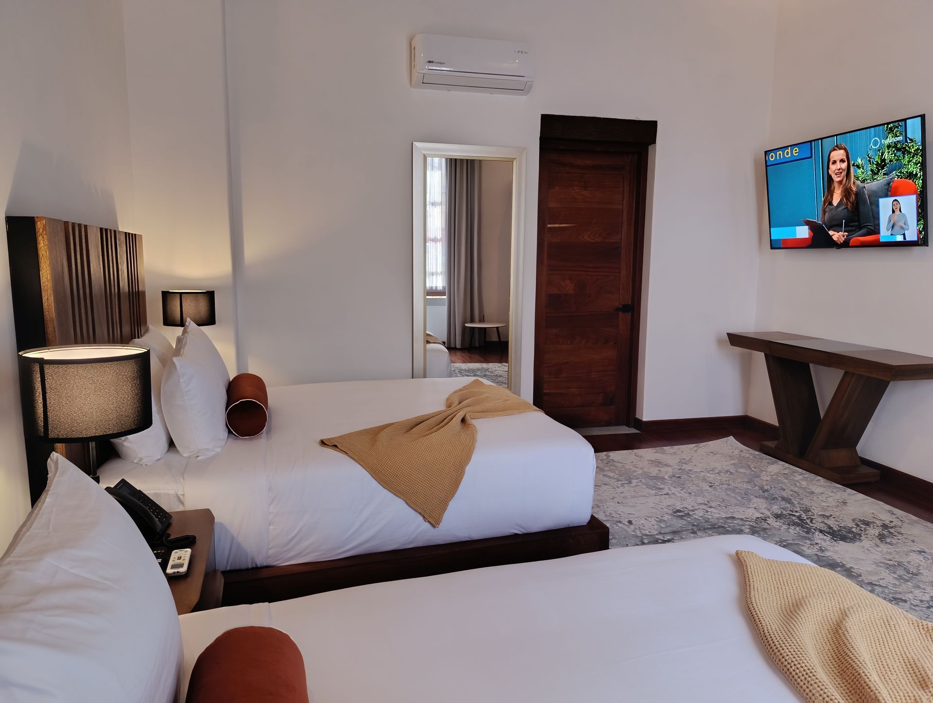 Habitación de hotel con dos camas, TV, puerta de madera y un espejo que refleja la entrada.