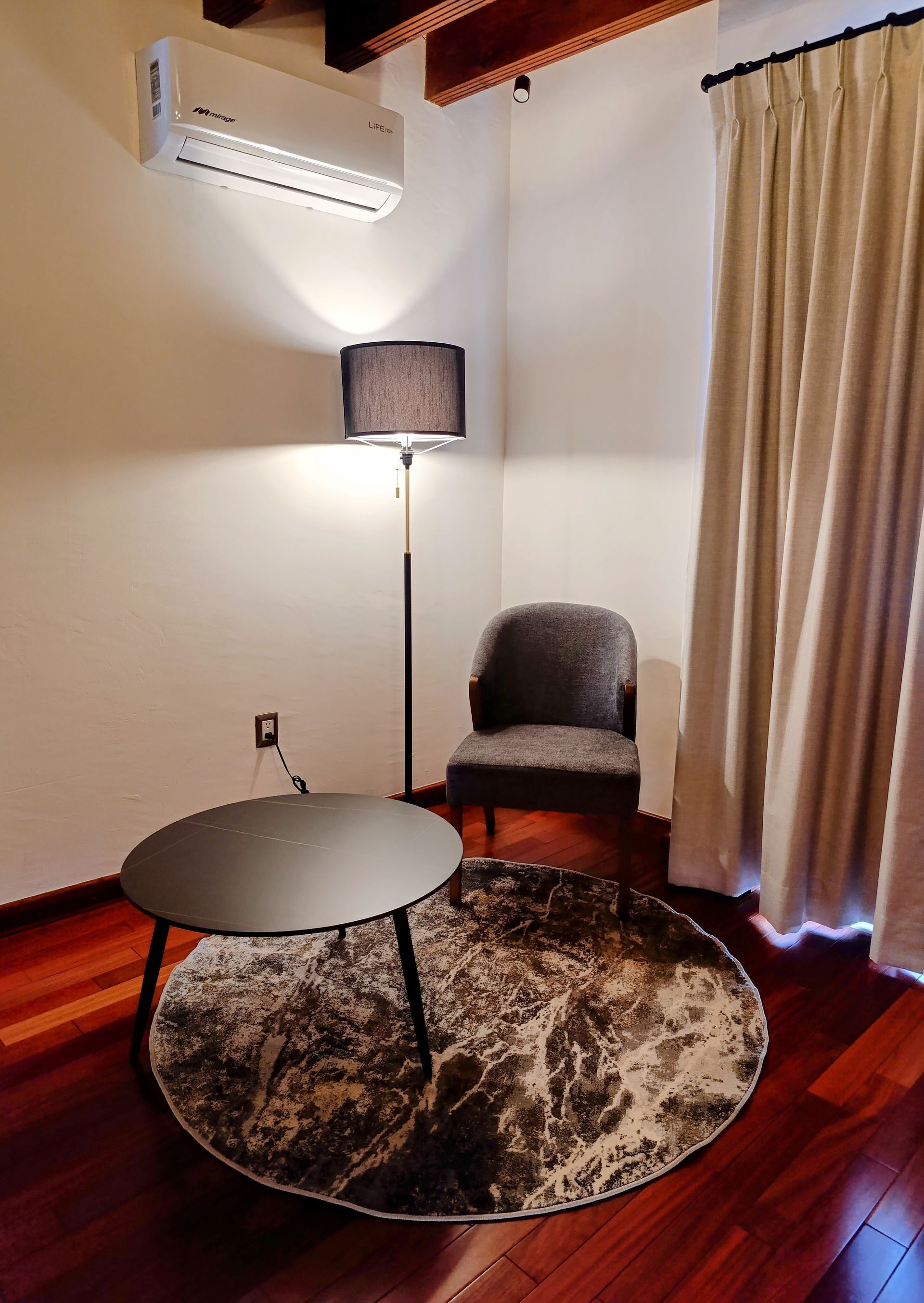 Rincón acogedor: silla, alfombra redonda, mesa pequeña, lámpara de pie, aire acondicionado y cortinas.