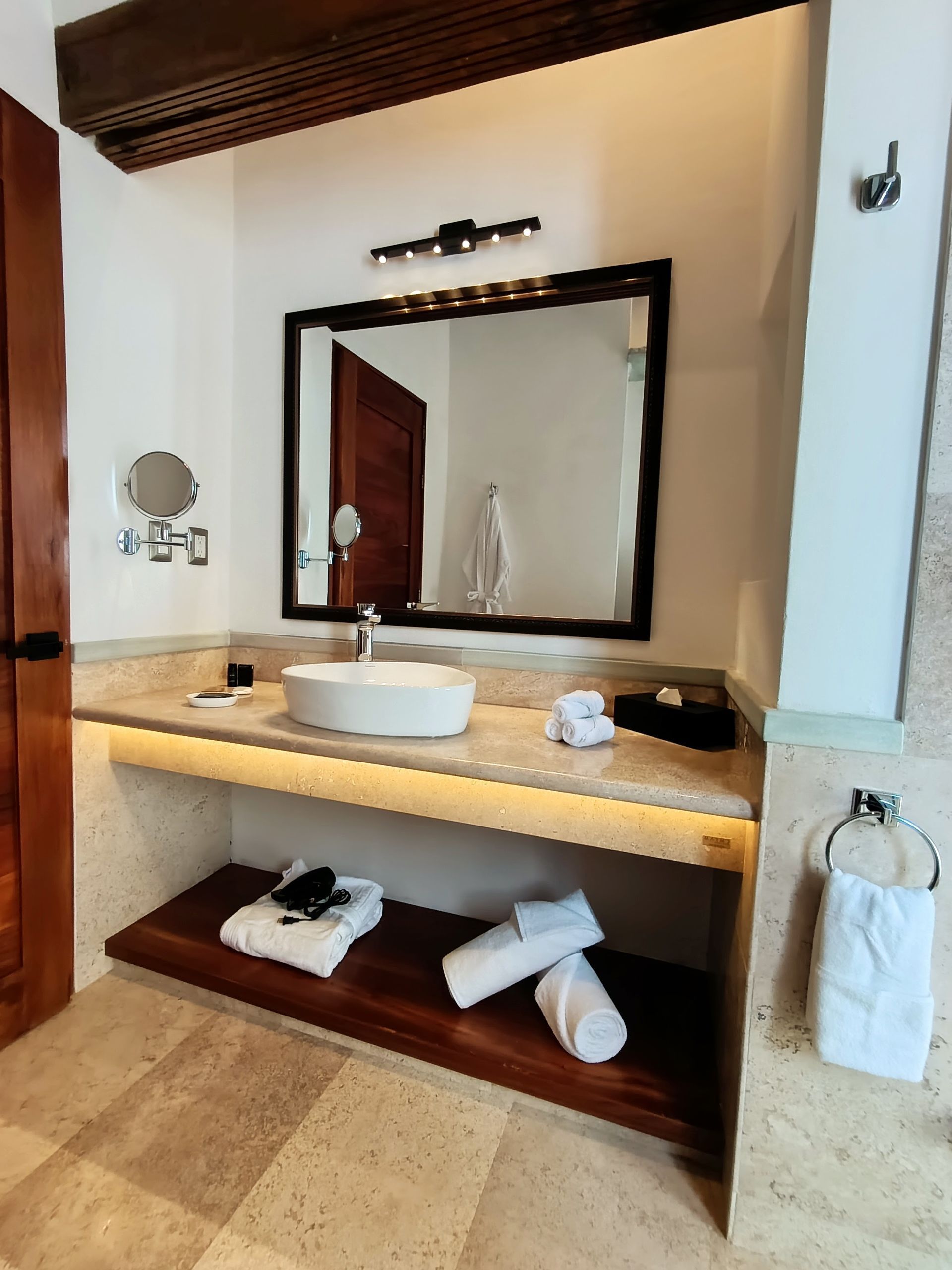 Baño con lavabo blanco, espejo grande y toallas en un estante de madera.