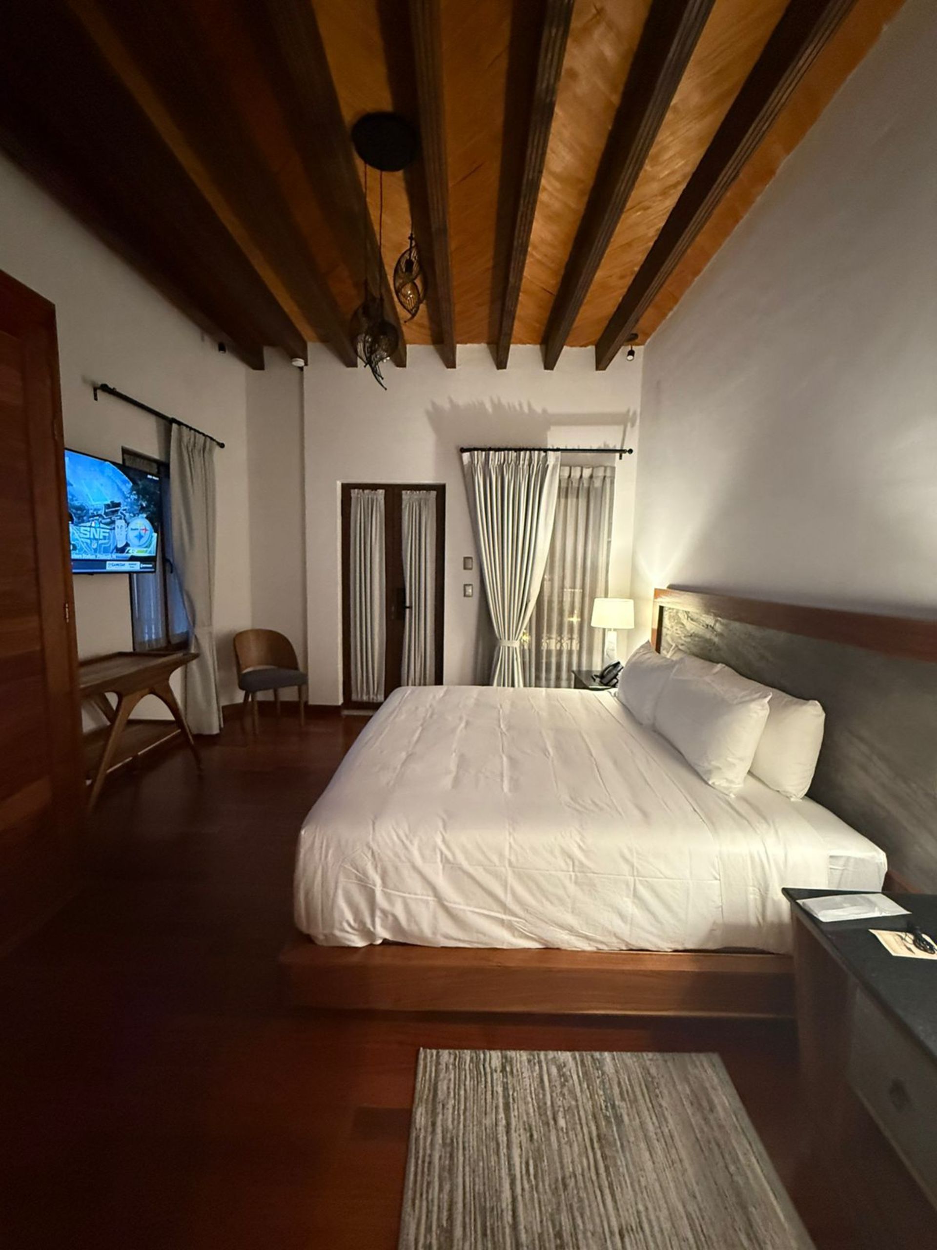 Dormitorio con vigas de madera en el techo, cama grande, TV y pisos de madera oscura.