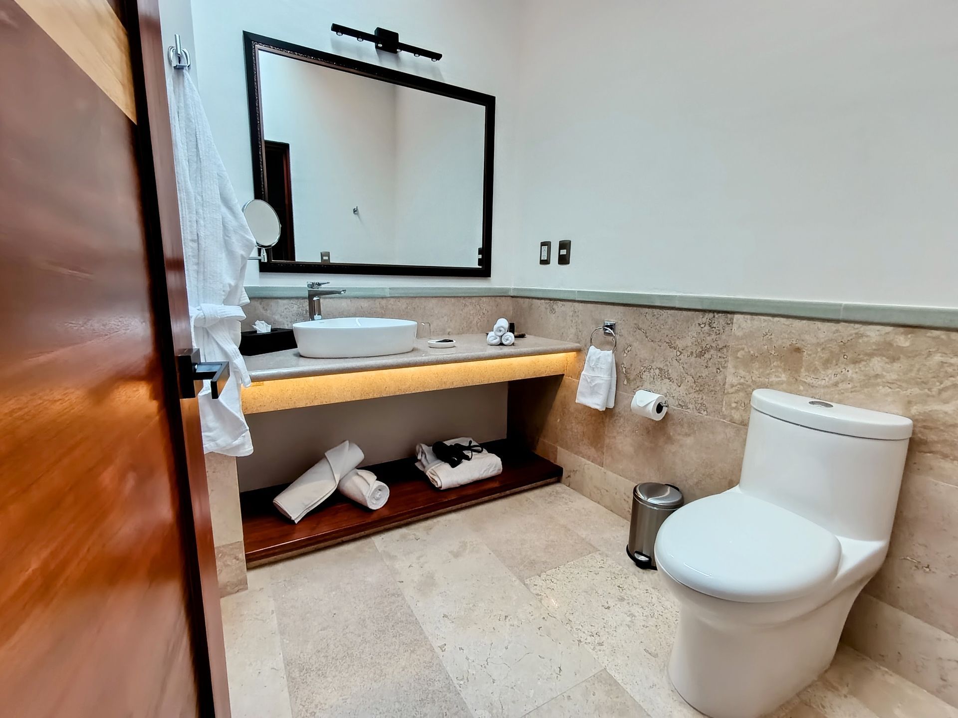 Baño con inodoro blanco, lavabo, espejo, toallas y puerta de madera. Paredes y suelo de piedra beige.