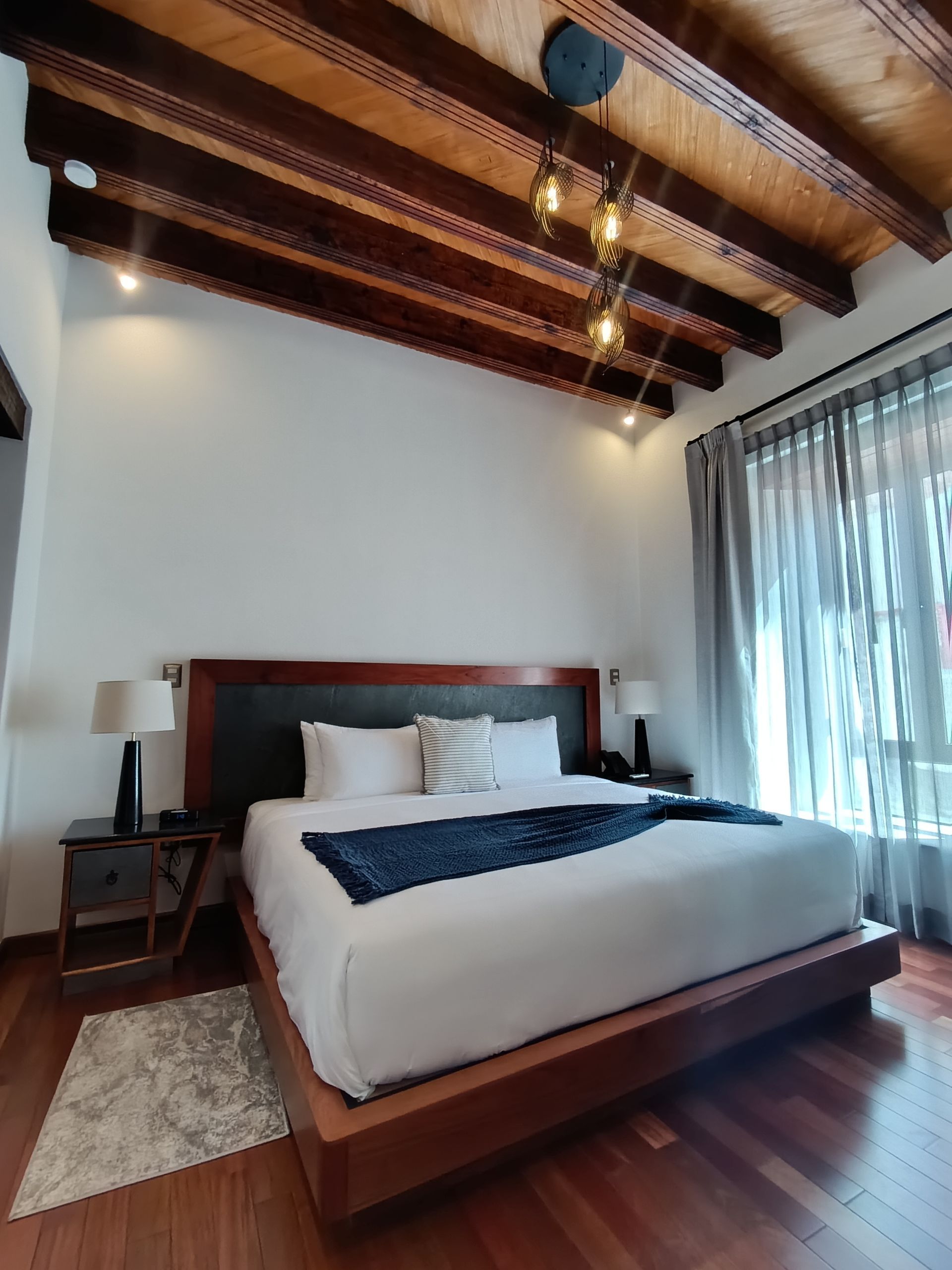 Dormitorio con vigas de madera en el techo, cama tamaño king y cortinas transparentes.