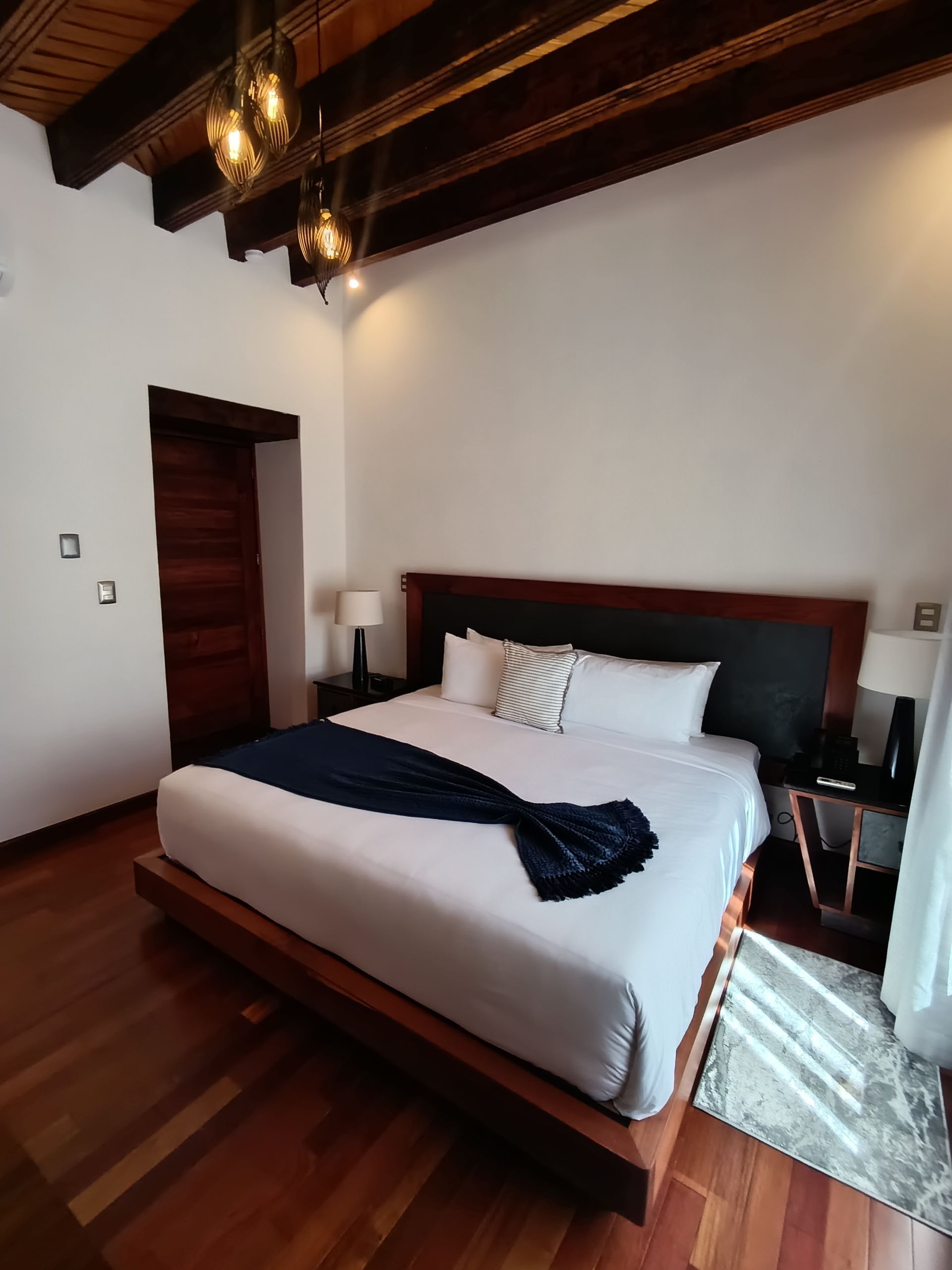 Dormitorio con cama tamaño king, detalles en madera oscura, paredes blancas y luces colgantes decorativas.