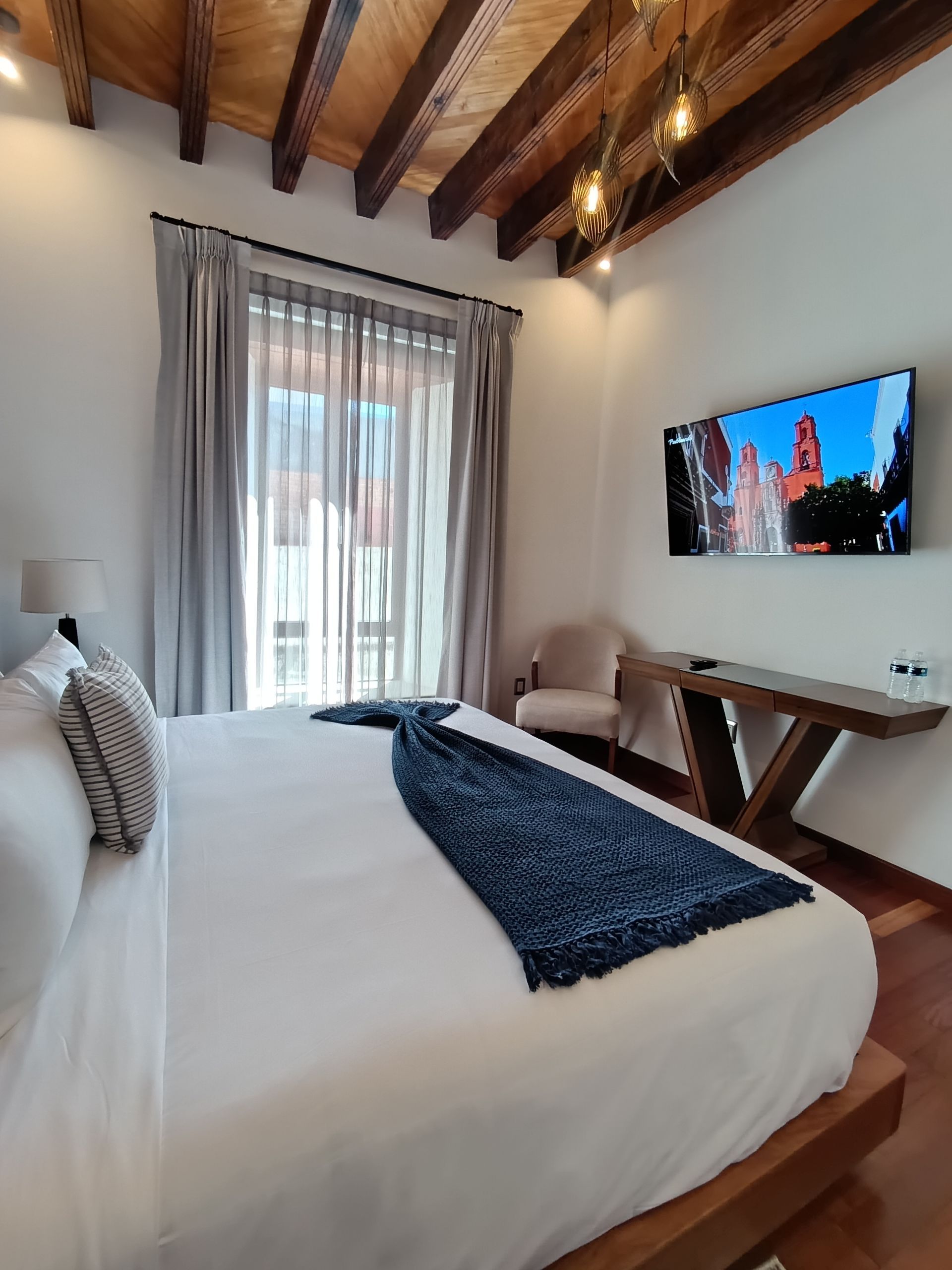 Dormitorio con cama grande, vigas de madera, TV y ventana con cortinas transparentes.