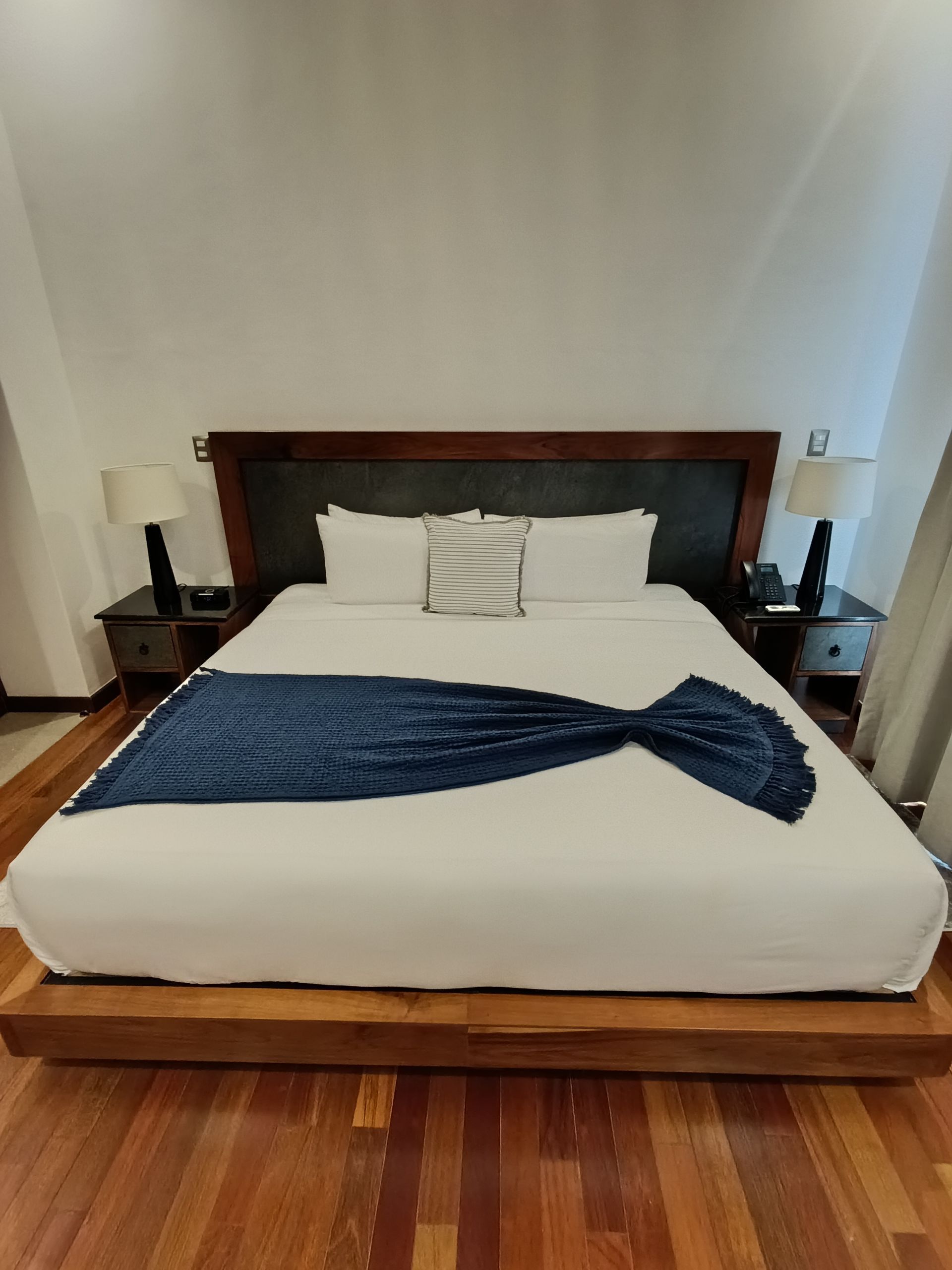 Dormitorio con cama grande, mesitas de noche, lámparas y suelo de madera. Manta azul sobre ropa de cama blanca.