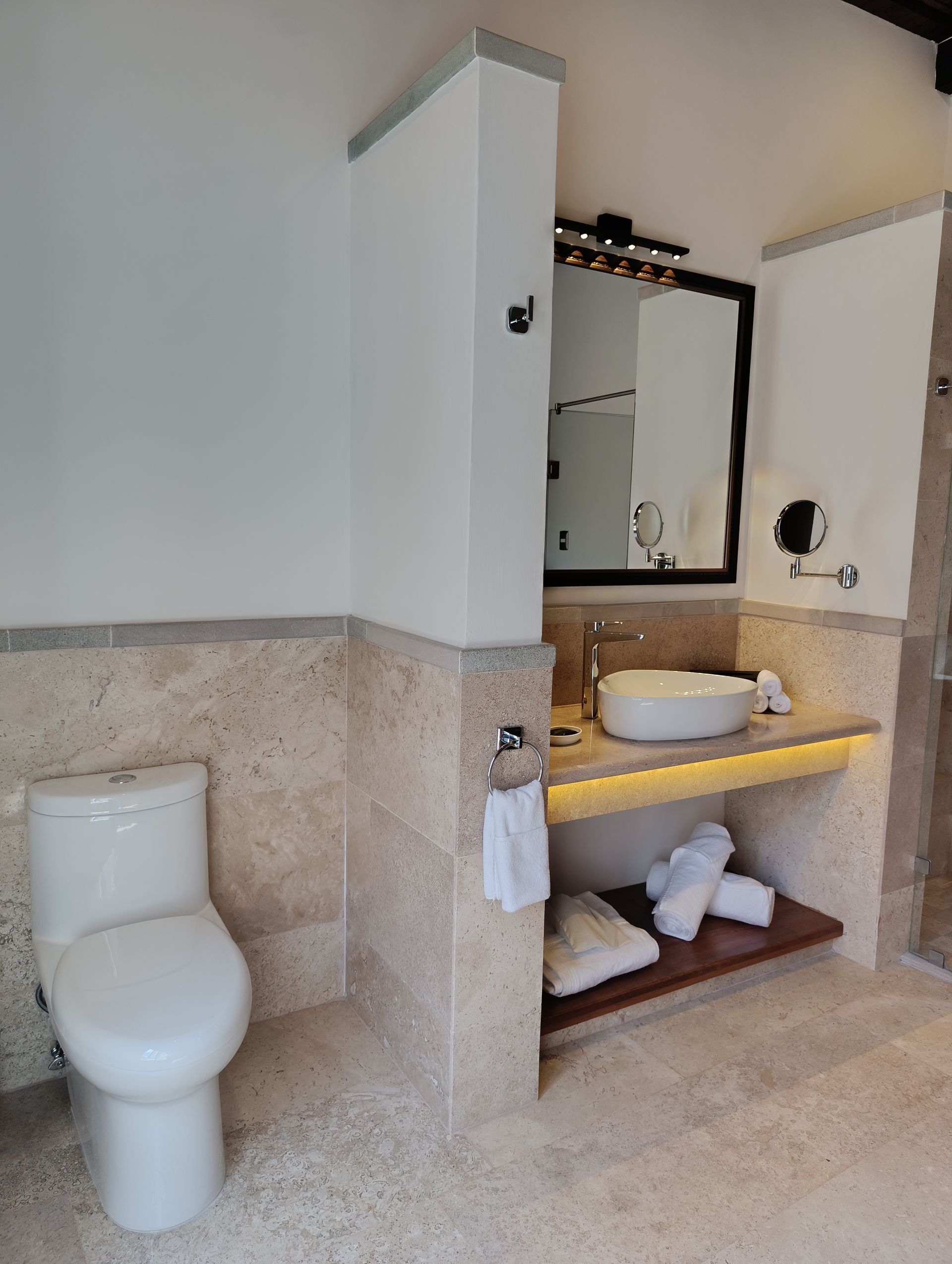 Baño con inodoro, lavabo, espejo y toallero. Paredes y suelo beige con detalles en blanco.