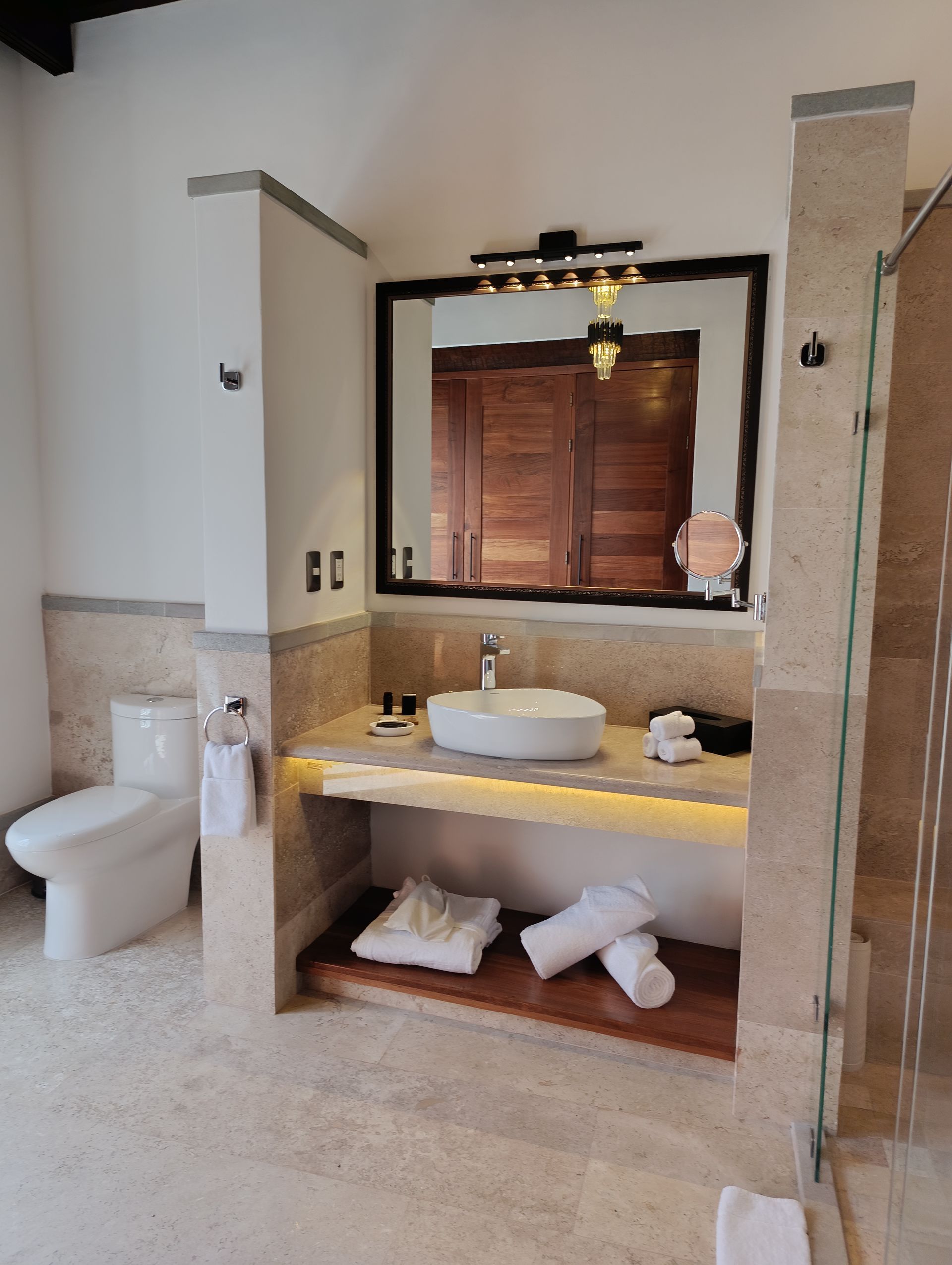 Baño con inodoro, lavabo y espejo blancos. Azulejos beige, detalles en madera y toallas.