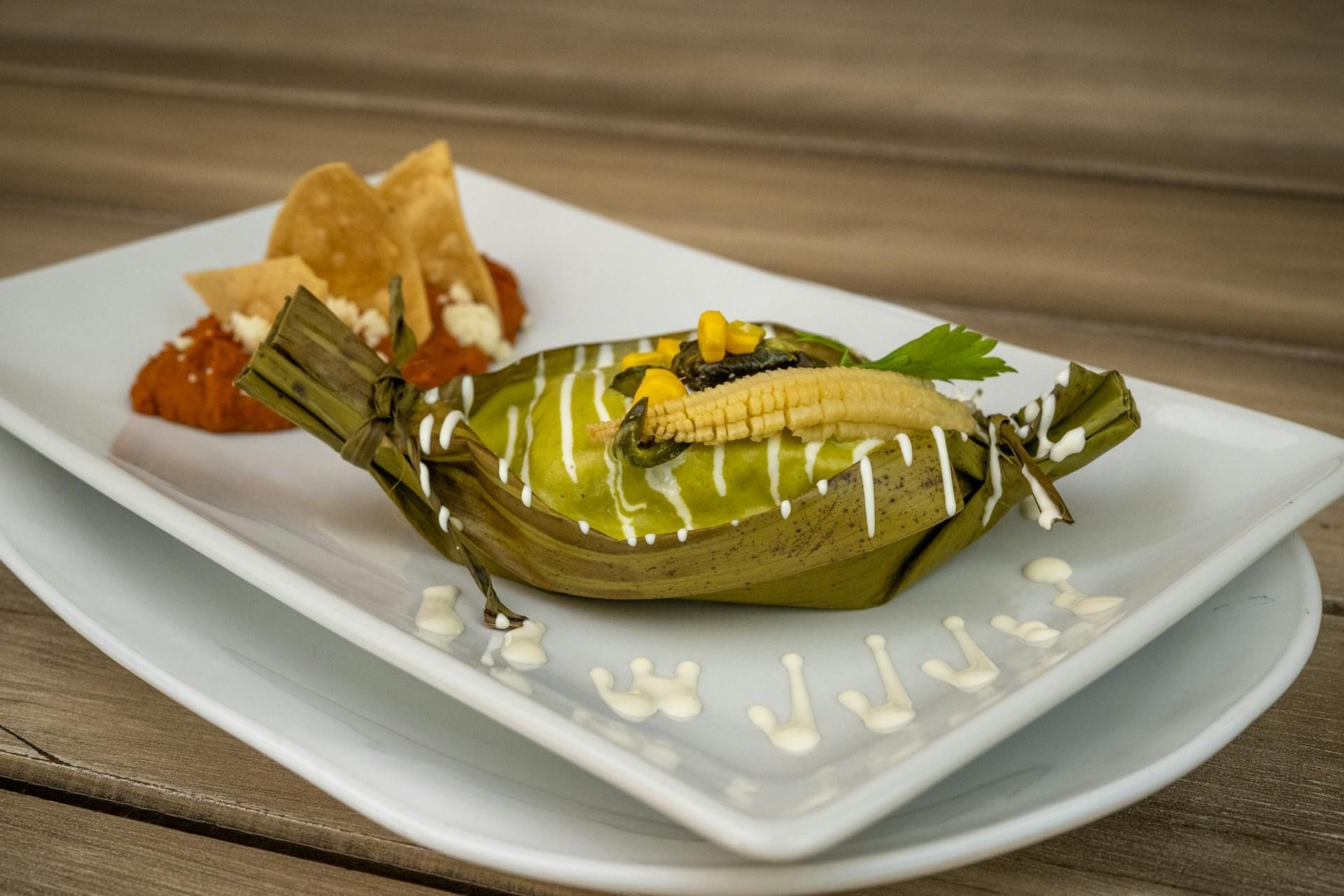 Tamal envuelto en hojas verdes con maíz, salsa y totopos en un plato blanco.