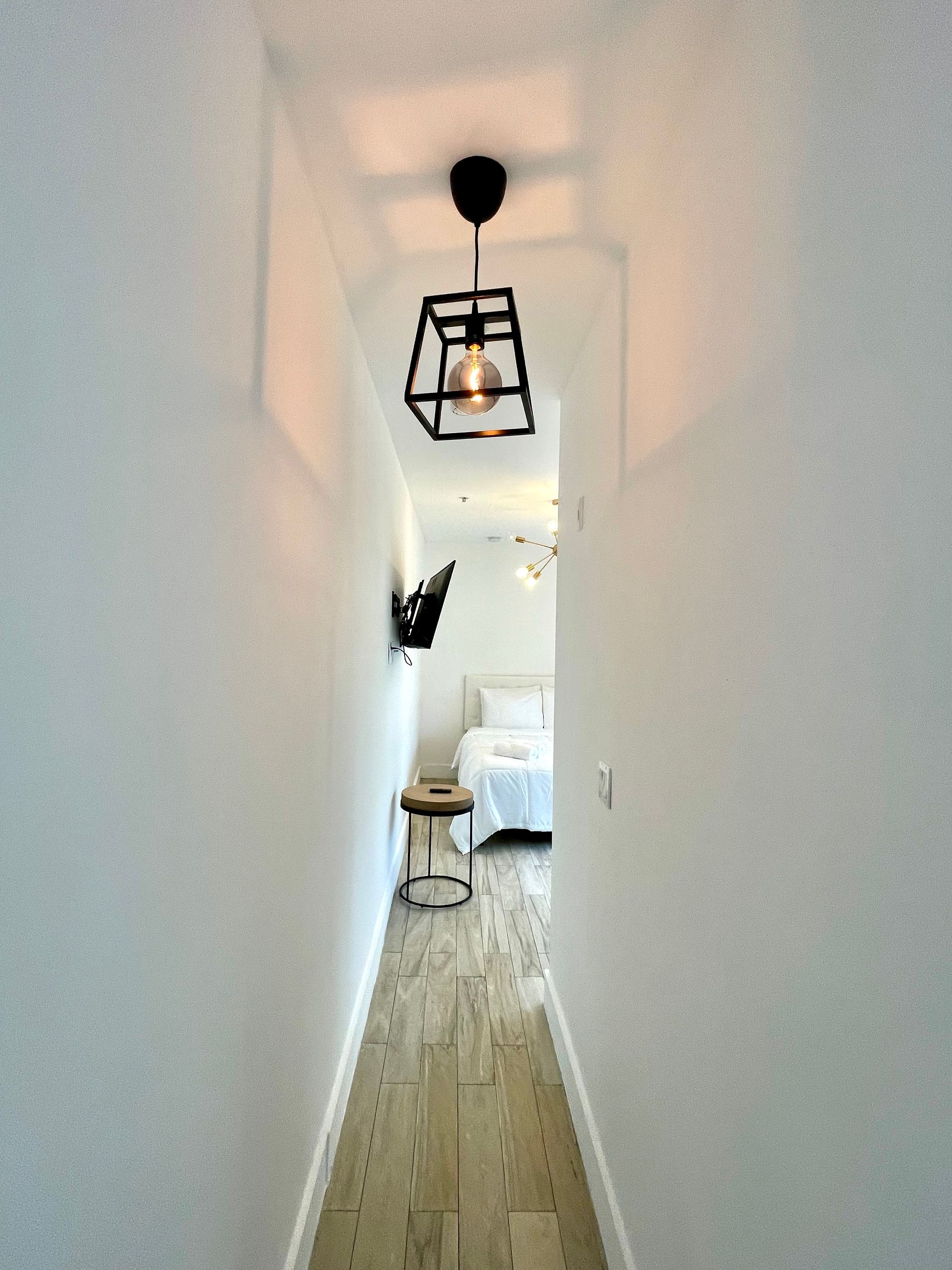 Narrow hallway with white walls and wooden floor, leading to a bedroom. A black geometric light hangs overhead.