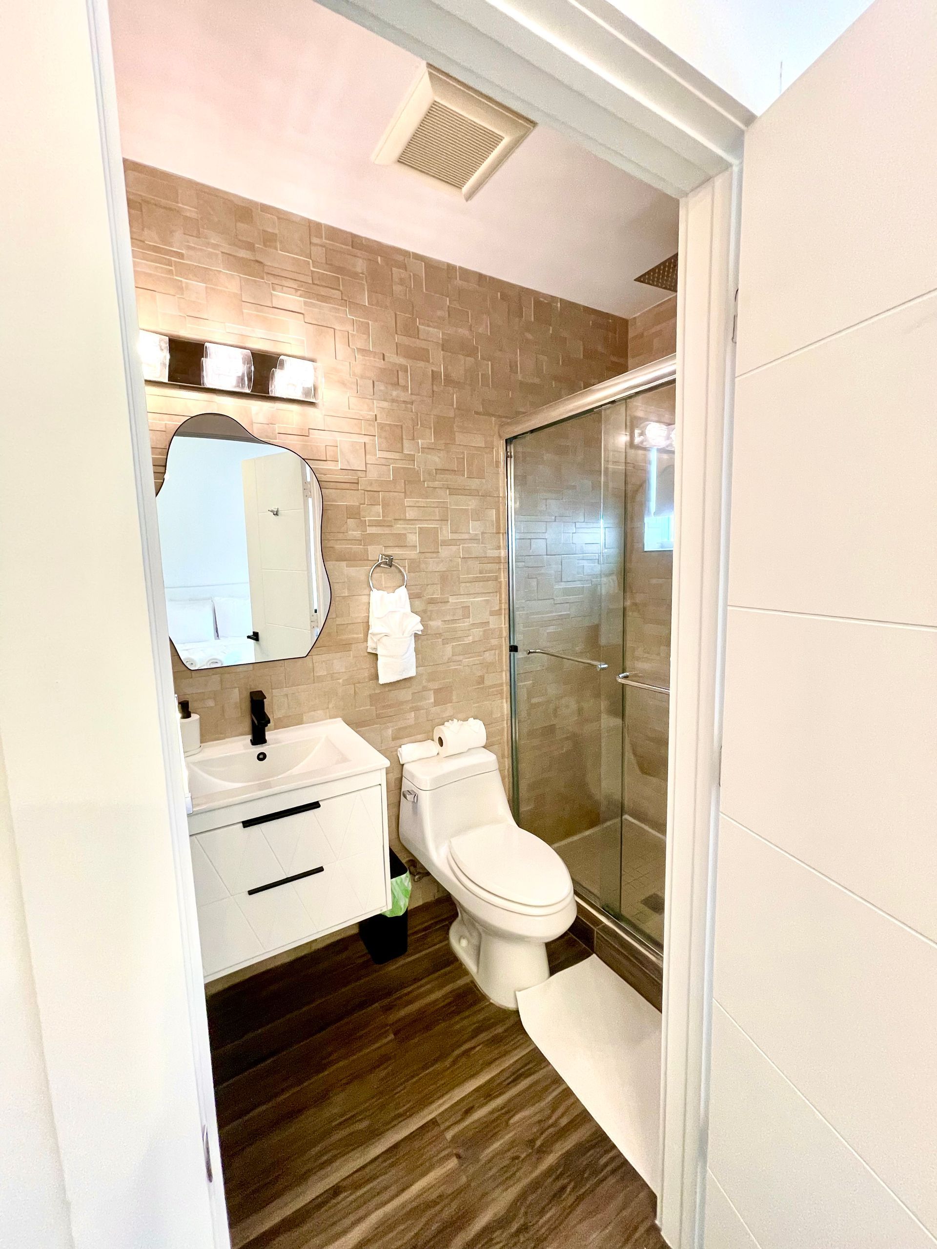 Bathroom with white vanity, toilet, and shower, brown tile wall and wooden floor.