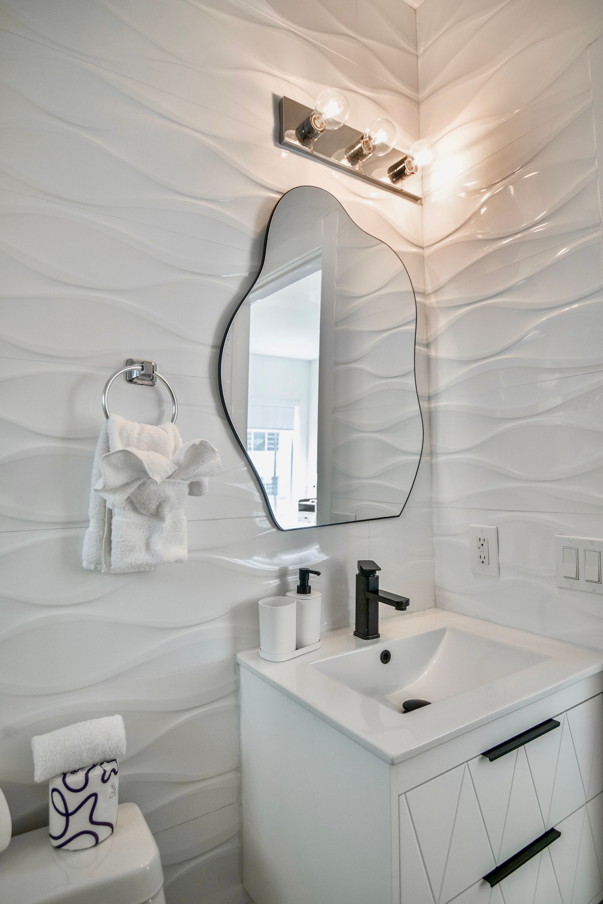 White bathroom with wavy tile, a unique mirror, and a vanity.
