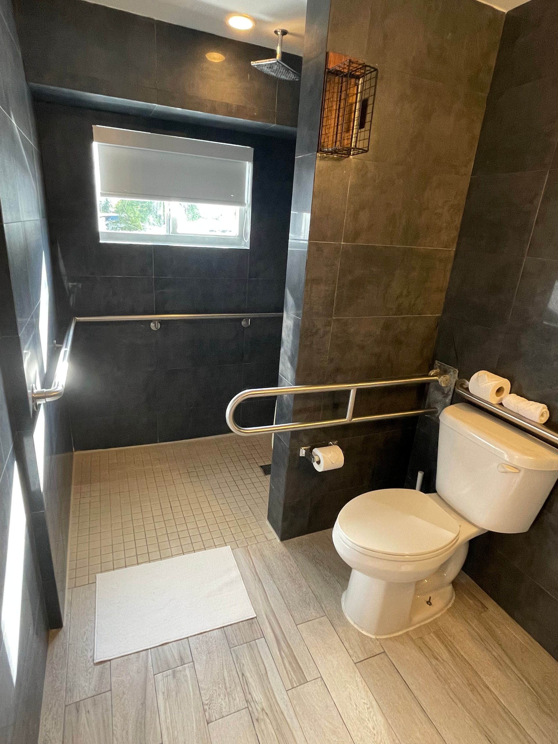 Accessible bathroom: gray tiled walls and shower, toilet with grab bars, window, and light wood-look floor.