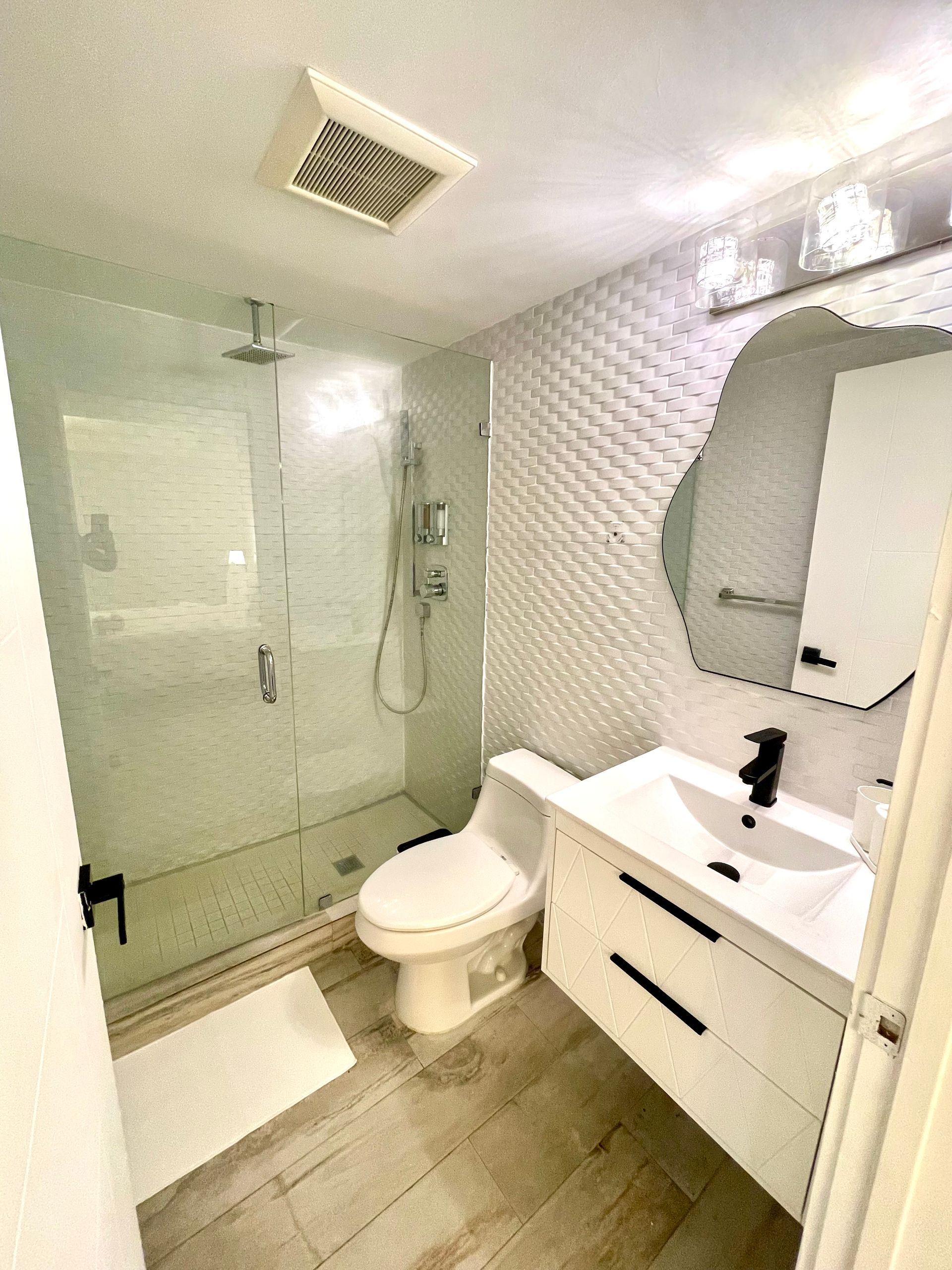 Modern bathroom with white vanity, patterned accent wall, glass shower, and light wood-look floor.