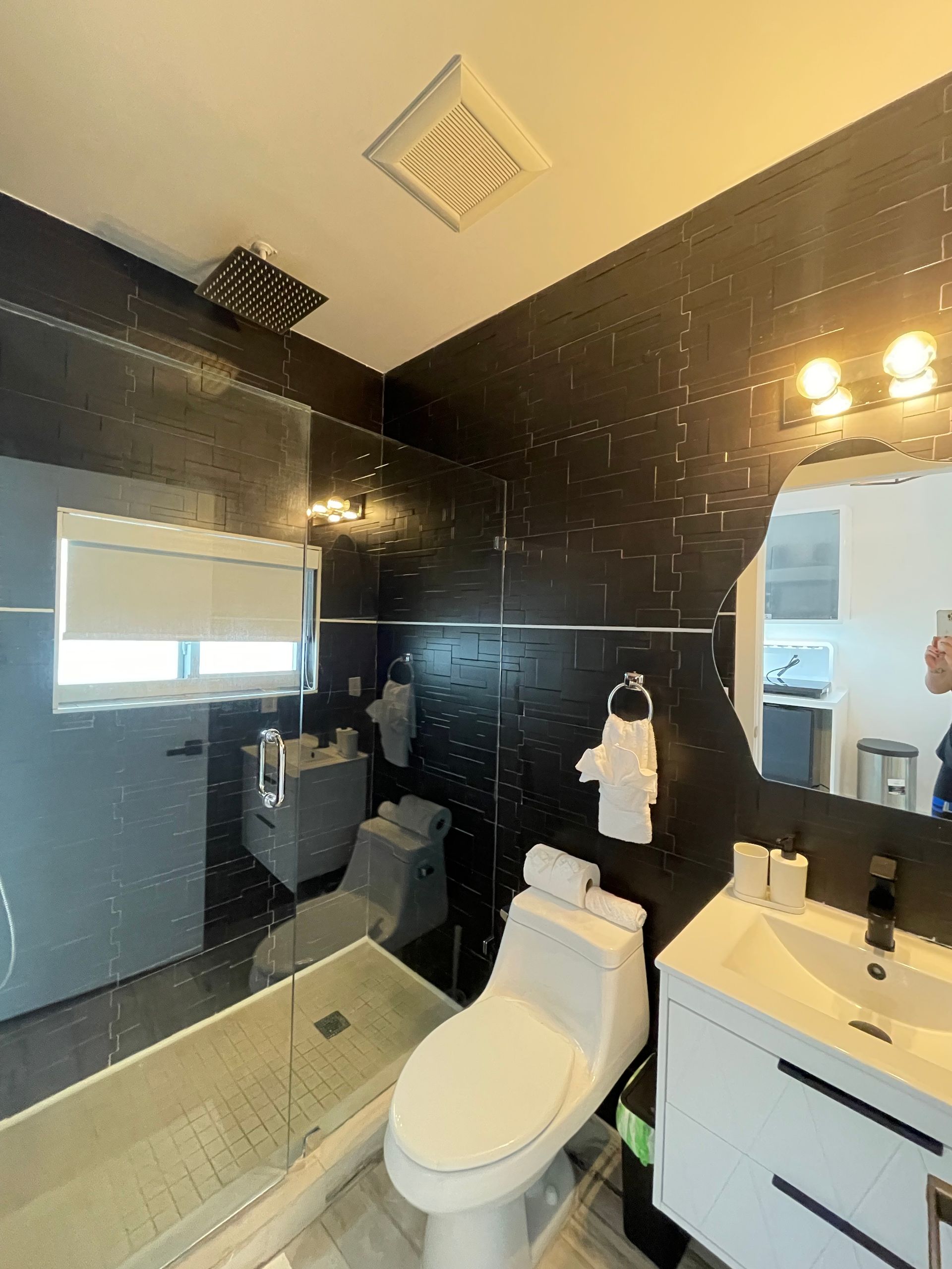 Modern bathroom with black tiled walls, toilet, sink, shower, and window.