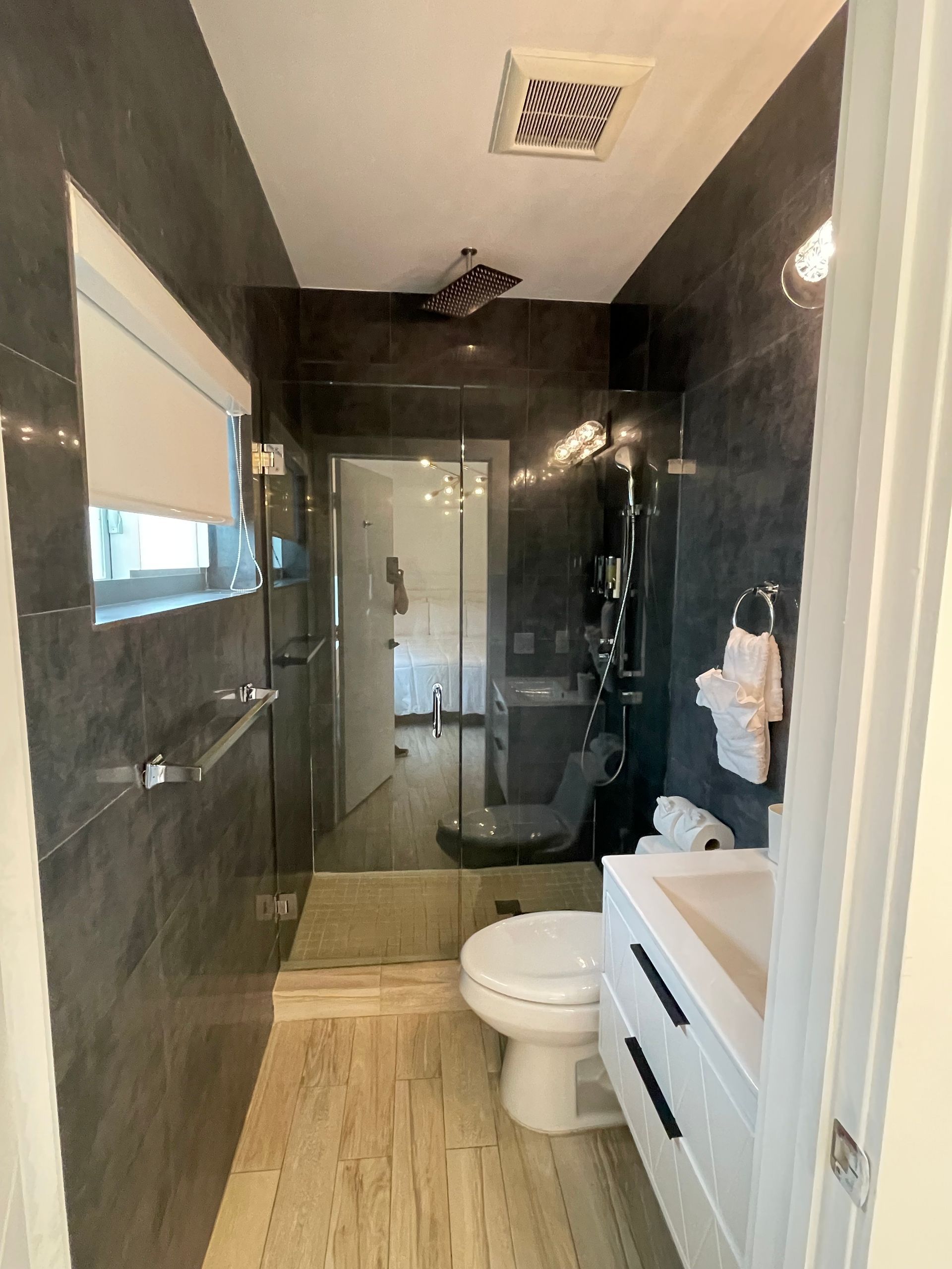 Narrow modern bathroom with dark gray tile walls, light wood-look floor, white vanity, and toilet.