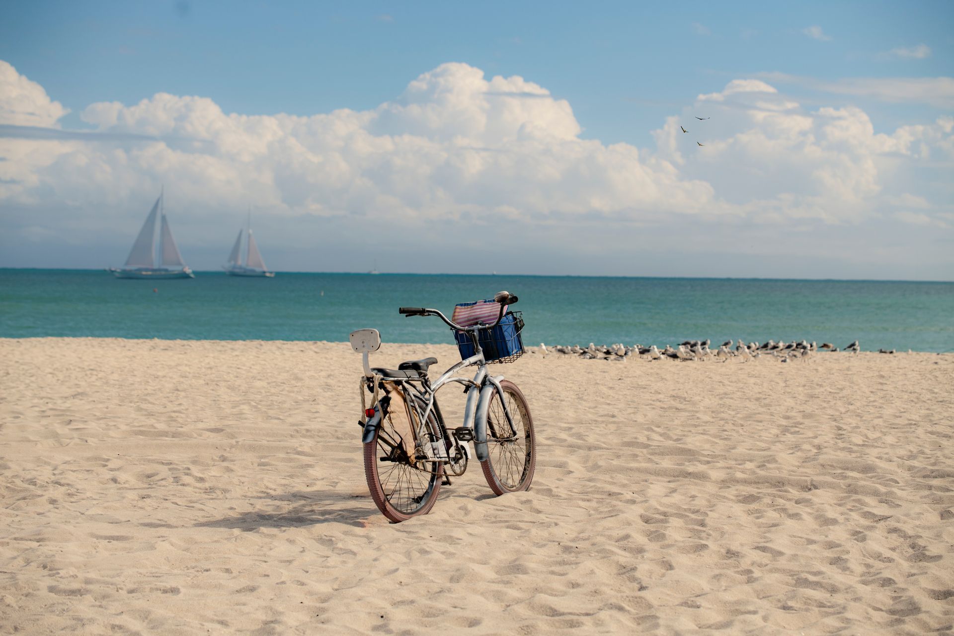 A bicycle on a sandy beach, turquoise water and sailboats in background, sunny day.