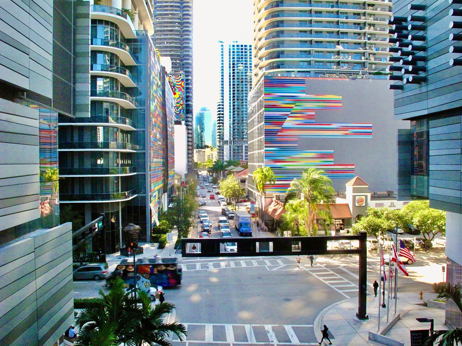 Street view of downtown Miami with high-rise buildings, cars, palm trees, and a colorful mural.