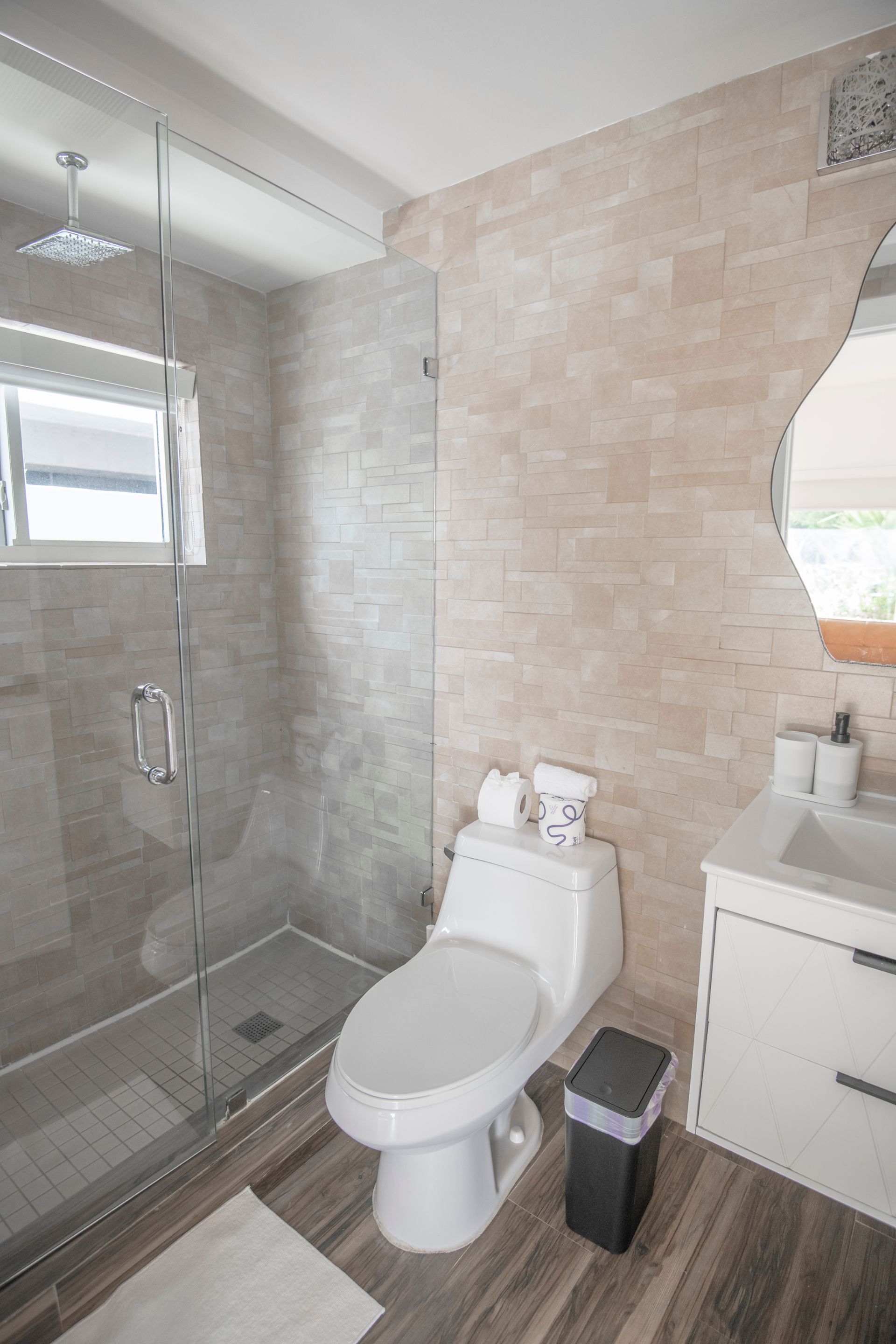 Modern bathroom with glass shower, toilet, vanity, and wood-look floors. Beige tiled walls.