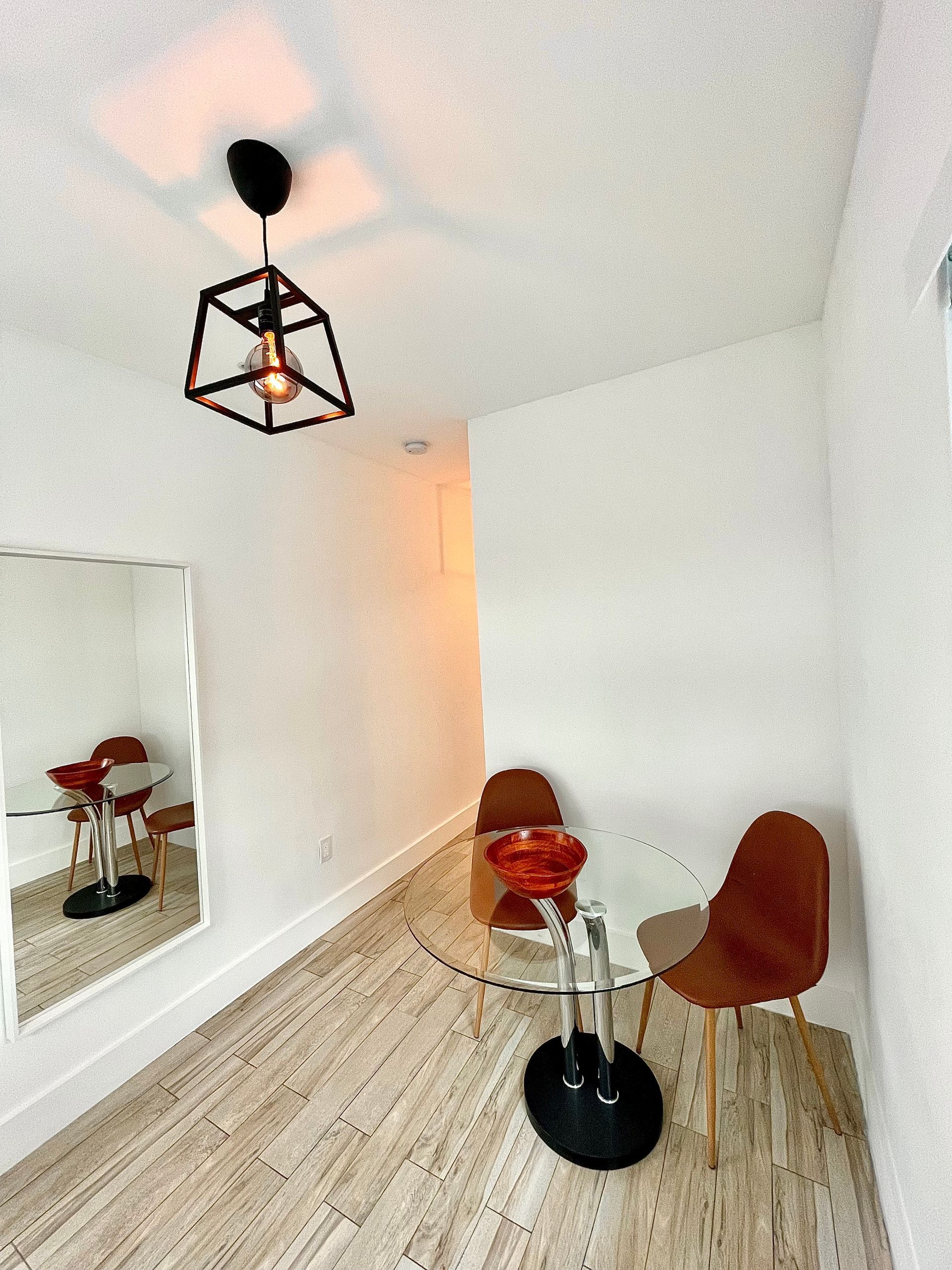 Small dining area with glass table, brown chairs, mirror, and black geometric light fixture.