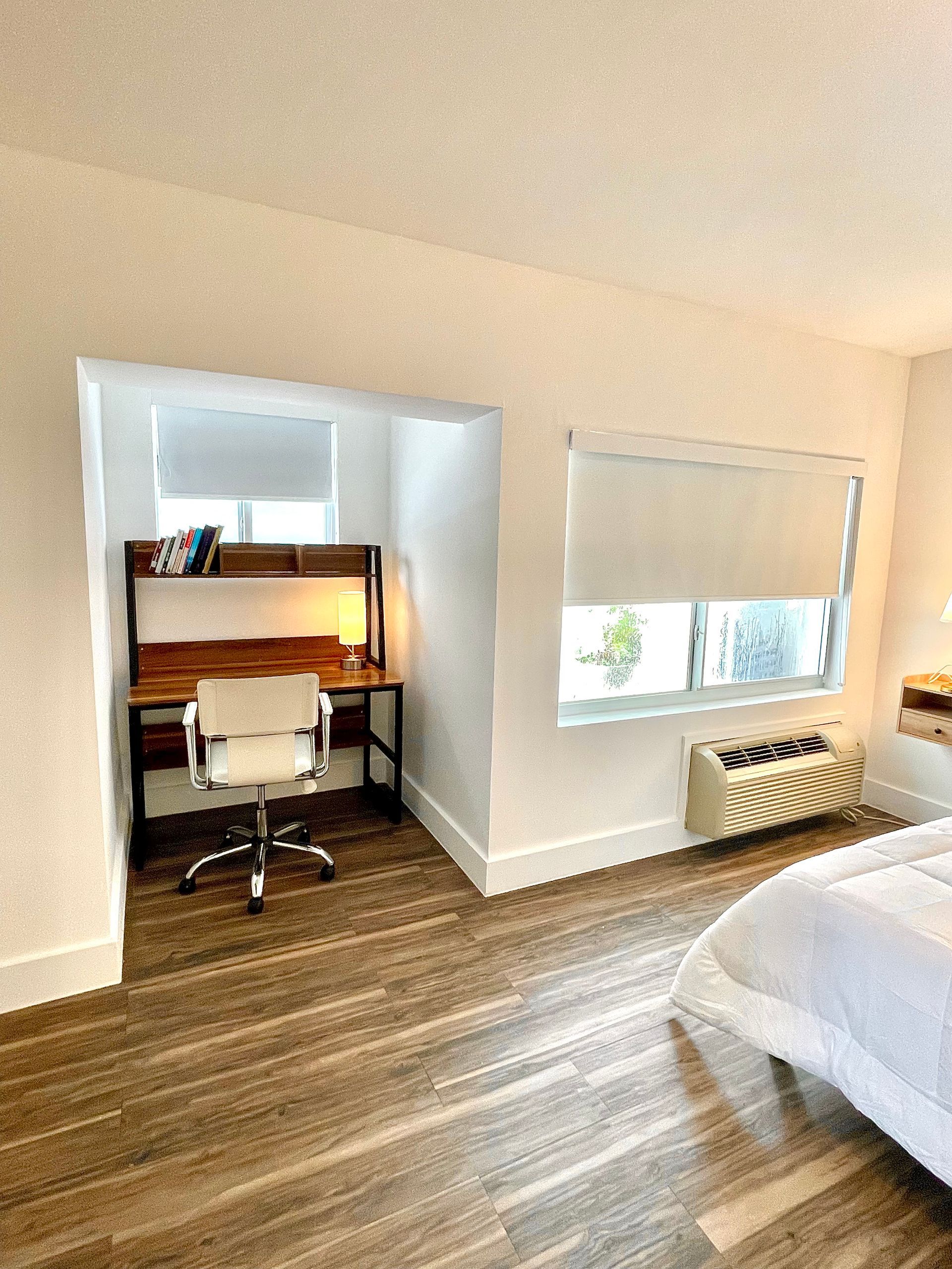 Built-in desk in hotel room with a desk chair, beneath a window, next to a bed. Brown wood floor.