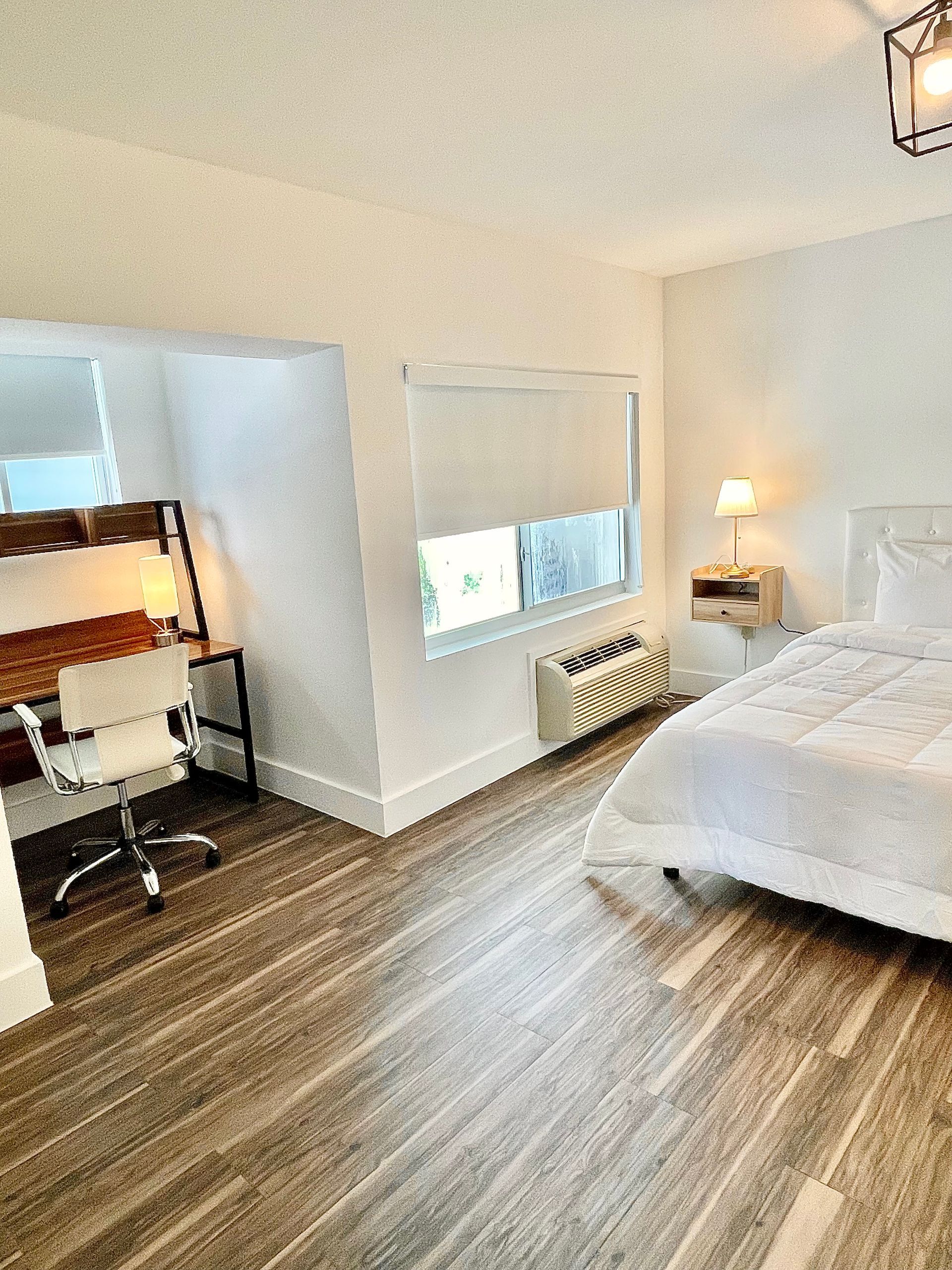 Bedroom with a desk, bed, and window. White walls, wood-look floor.  Desk lamp on.