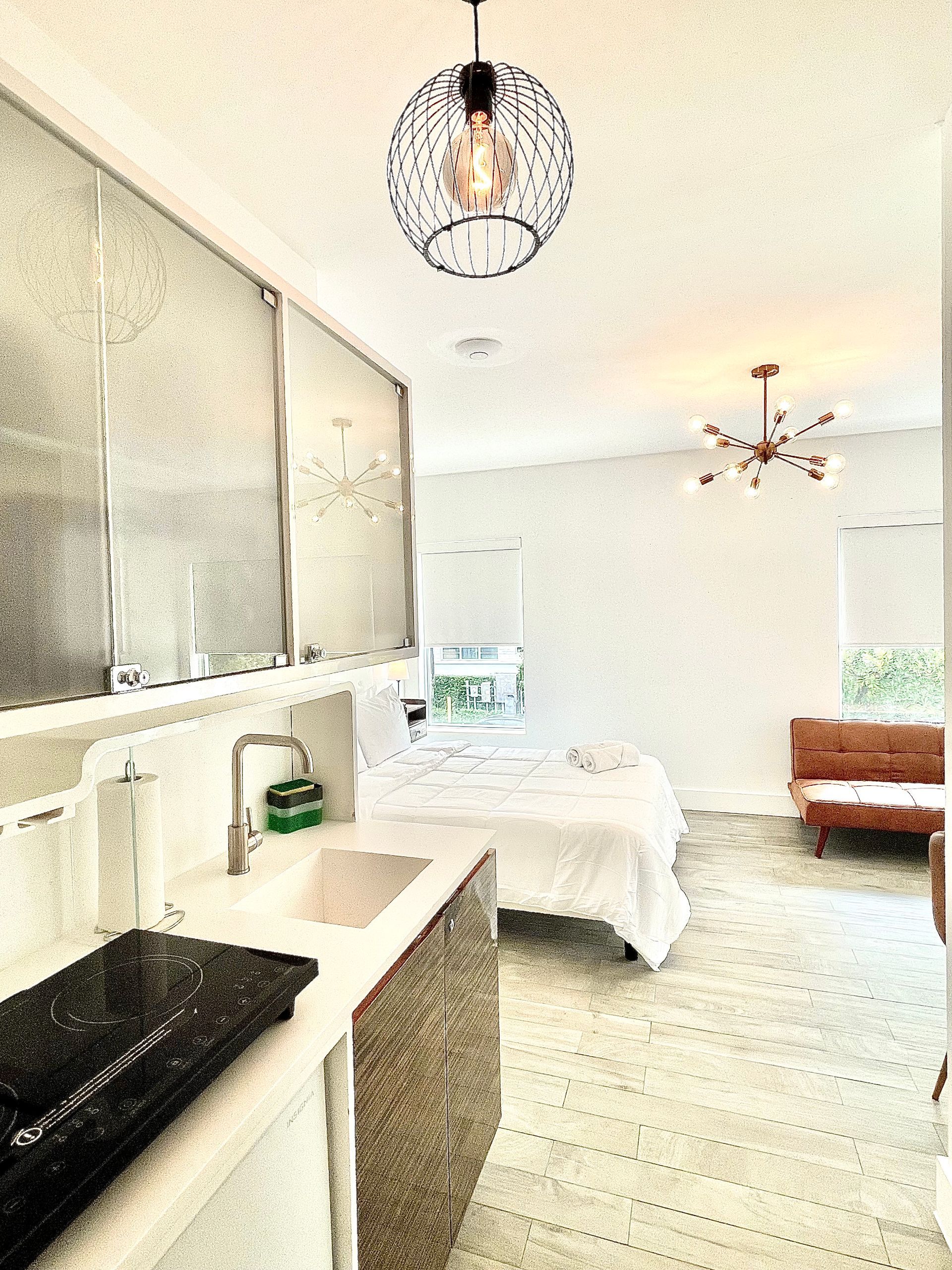 A small, bright apartment with a kitchenette, bed, and mid-century modern furniture; a hanging orb light.
