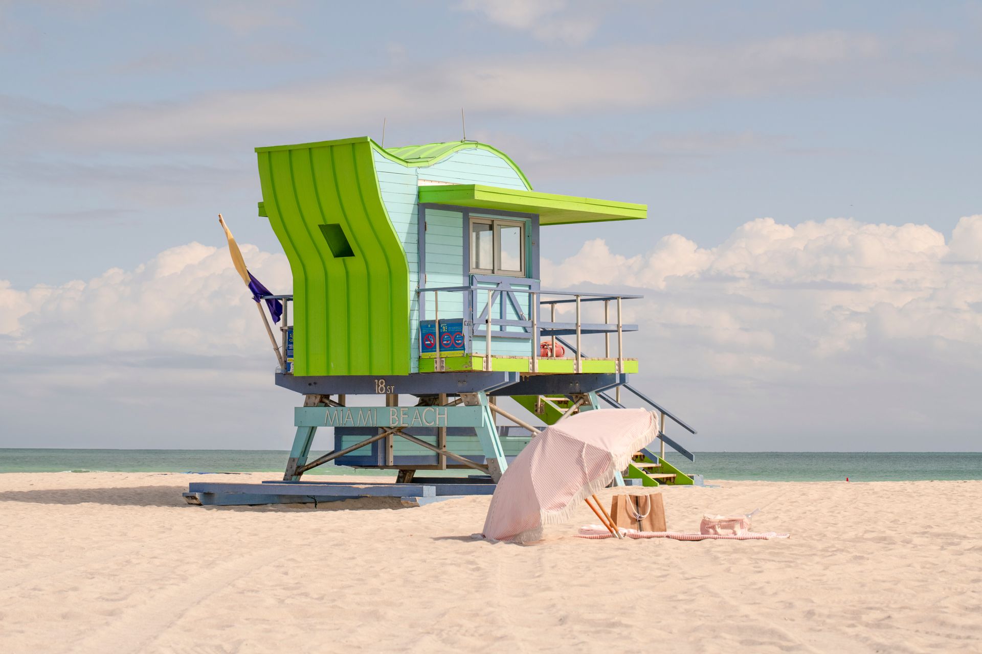 Bright green lifeguard stand on a white sandy beach with a pink umbrella.