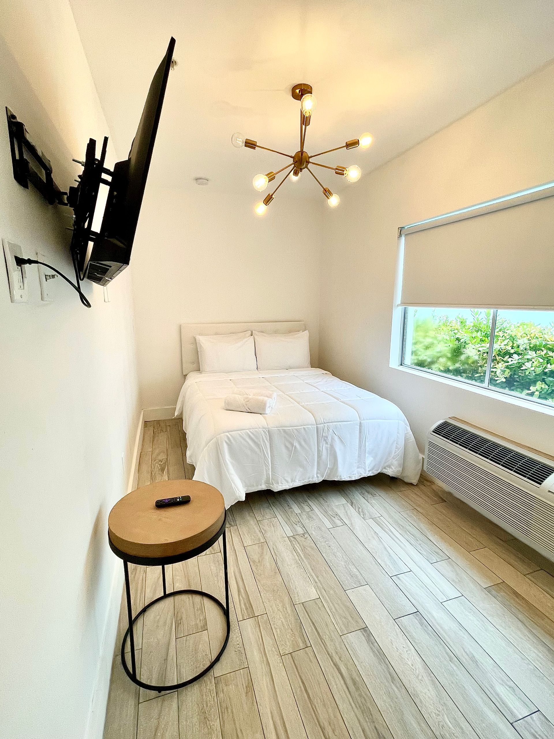 Small bedroom with white walls, bed, TV, and a round side table. Bright lighting.