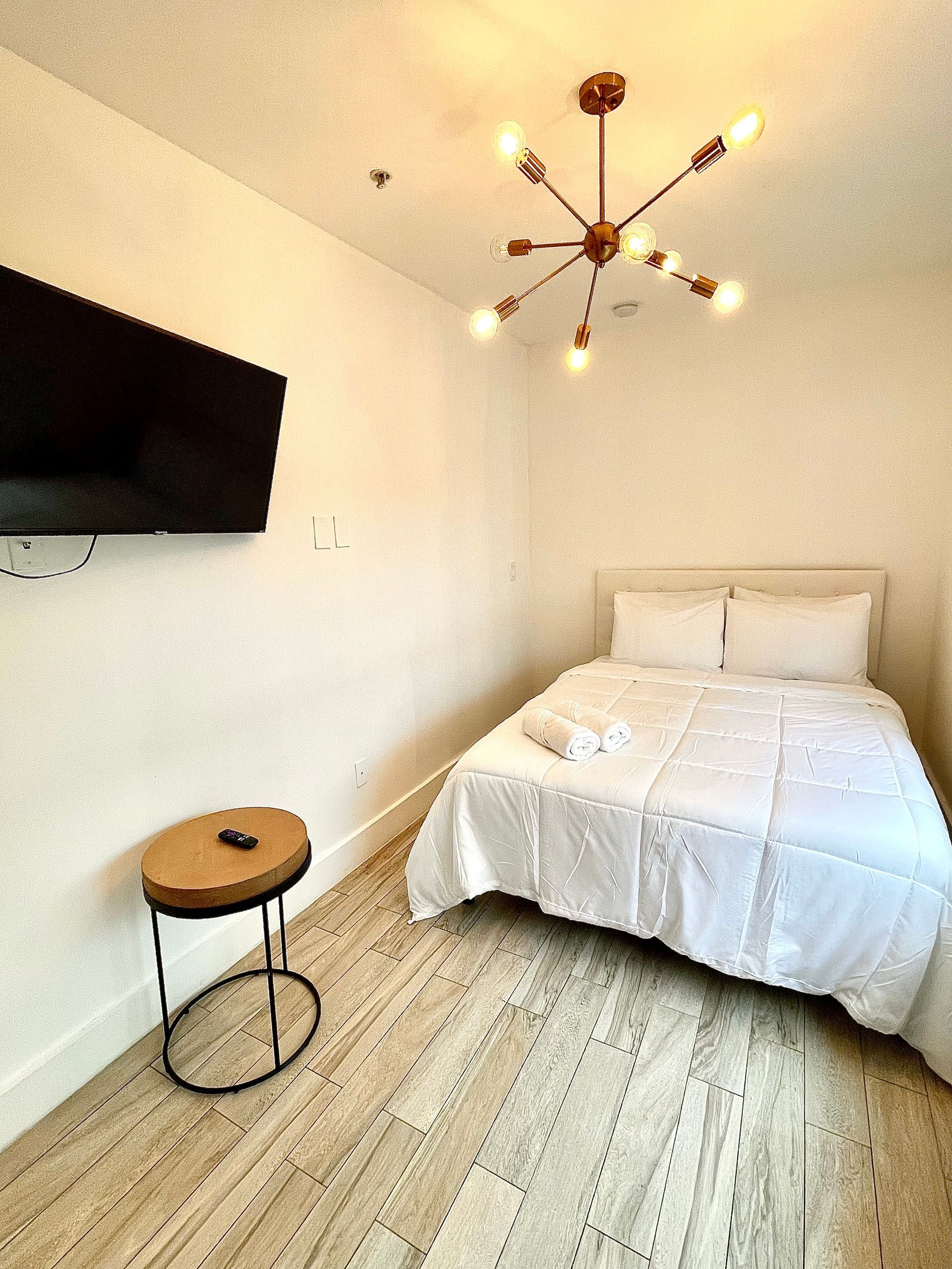 Small, simple bedroom with a bed, TV, side table, and a unique light fixture.