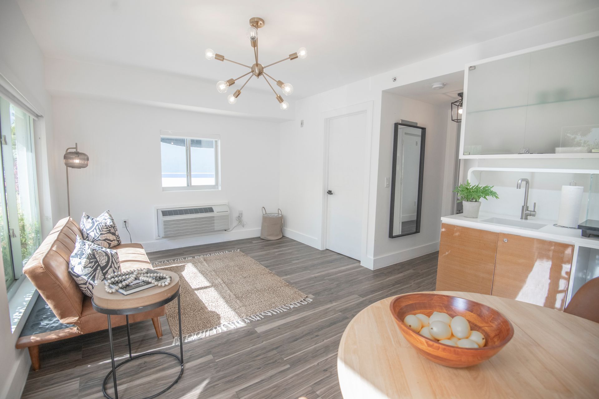 Bright living space with a brown leather sofa, wooden table, and a small kitchen area.