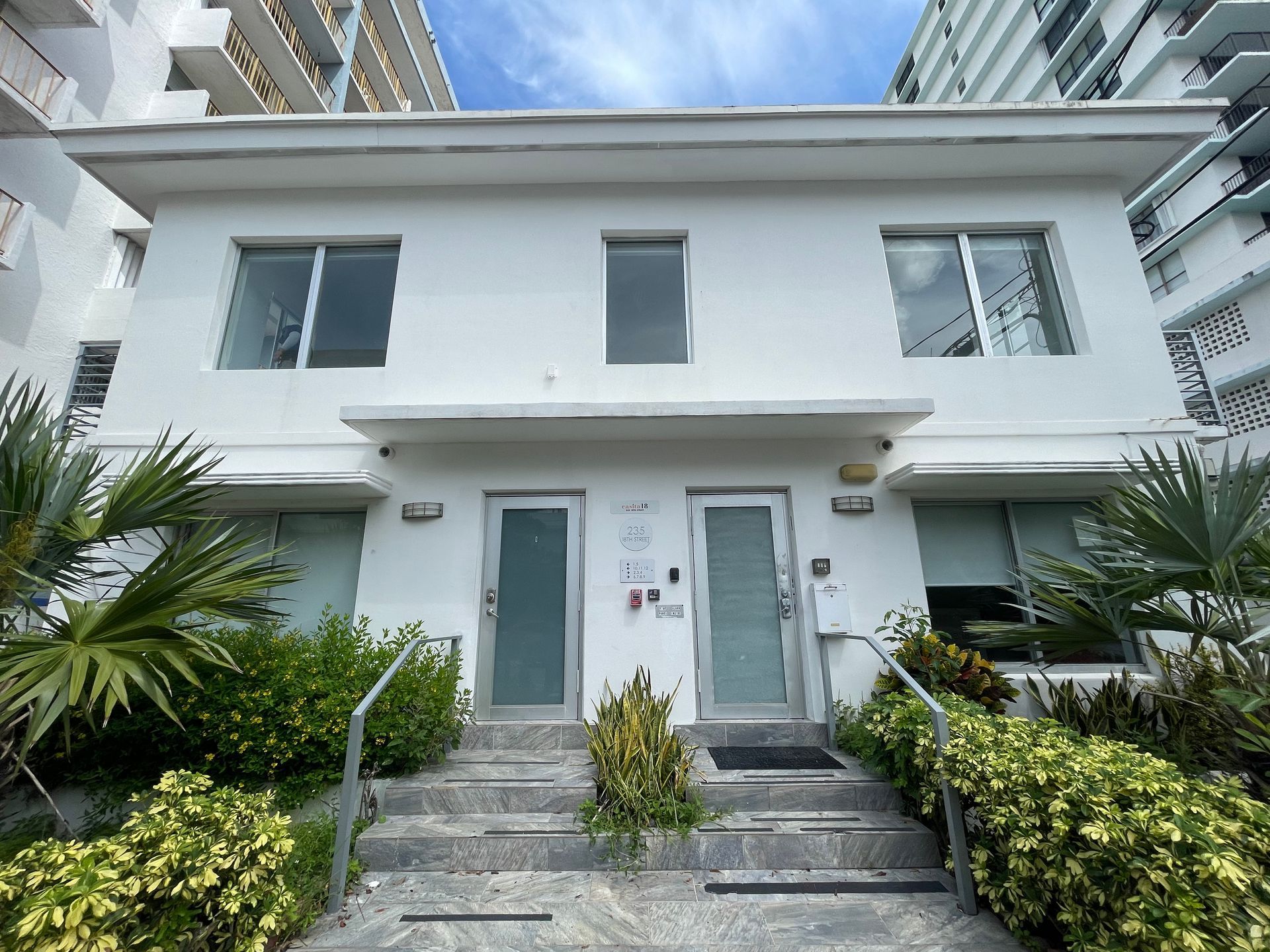 White two-story building with front doors, windows, and steps. Green bushes line the building.