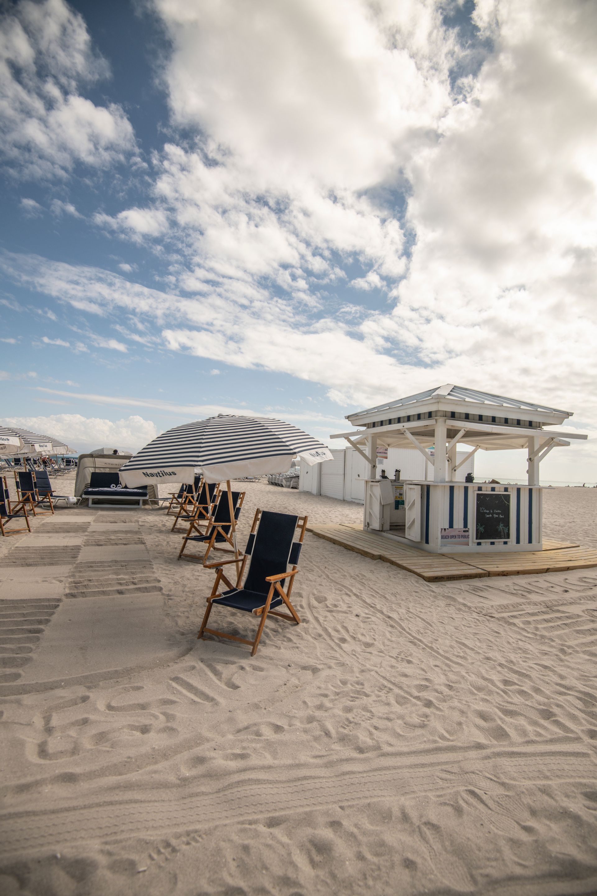Beach scene with chairs, striped umbrellas, and a white bar under a cloudy sky.