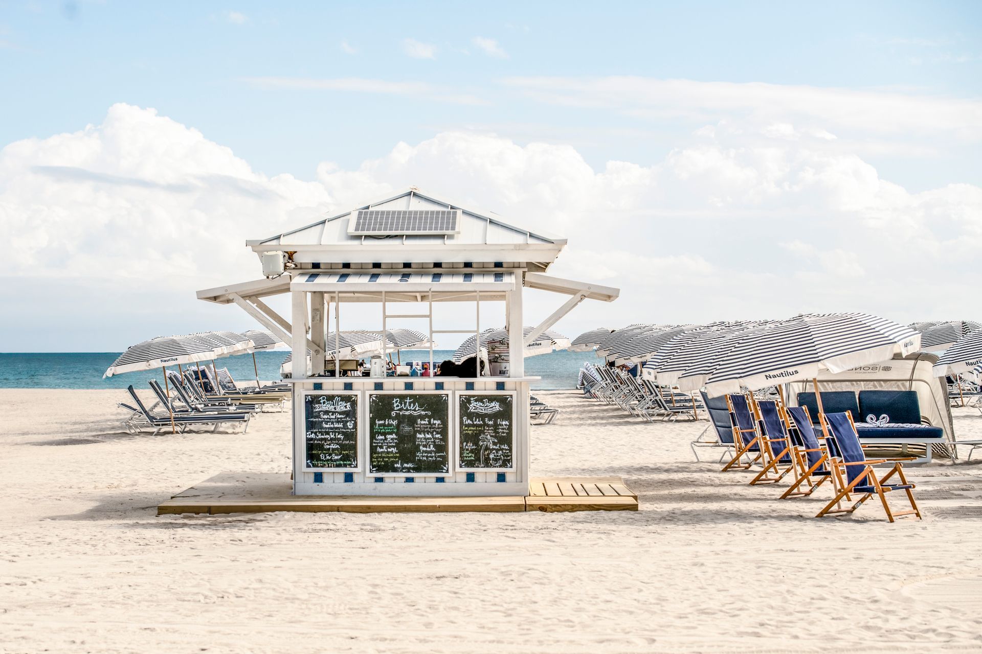 White beach bar with menu on a sunny beach with blue water, chairs, and umbrellas.