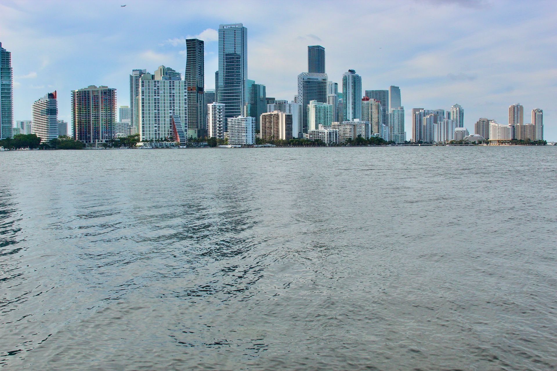 City skyline with many high-rise buildings on the horizon, across a body of water under a cloudy sky.