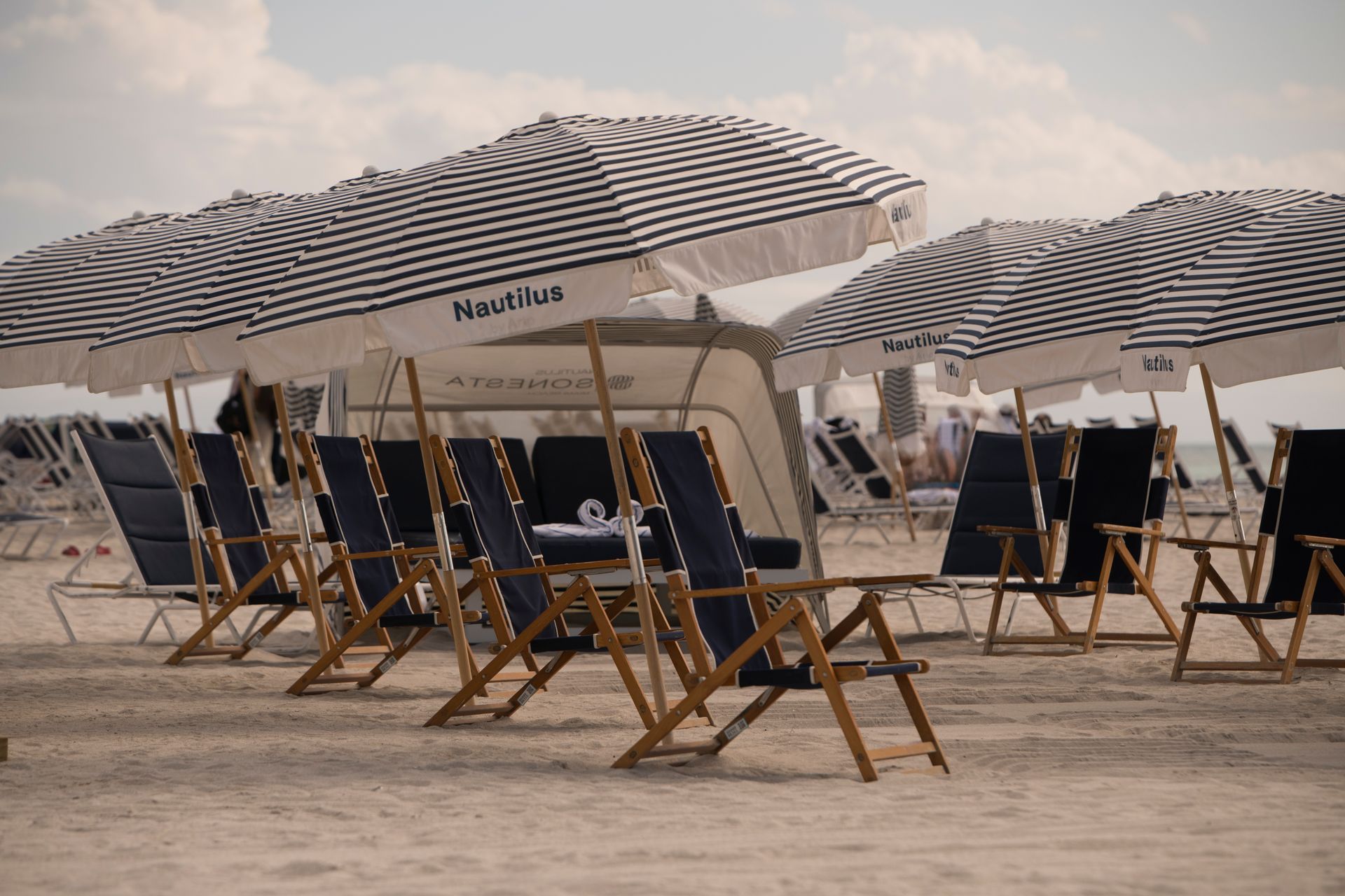 Beach chairs and umbrellas on the sand, with the Nautilus logo, under a cloudy sky.