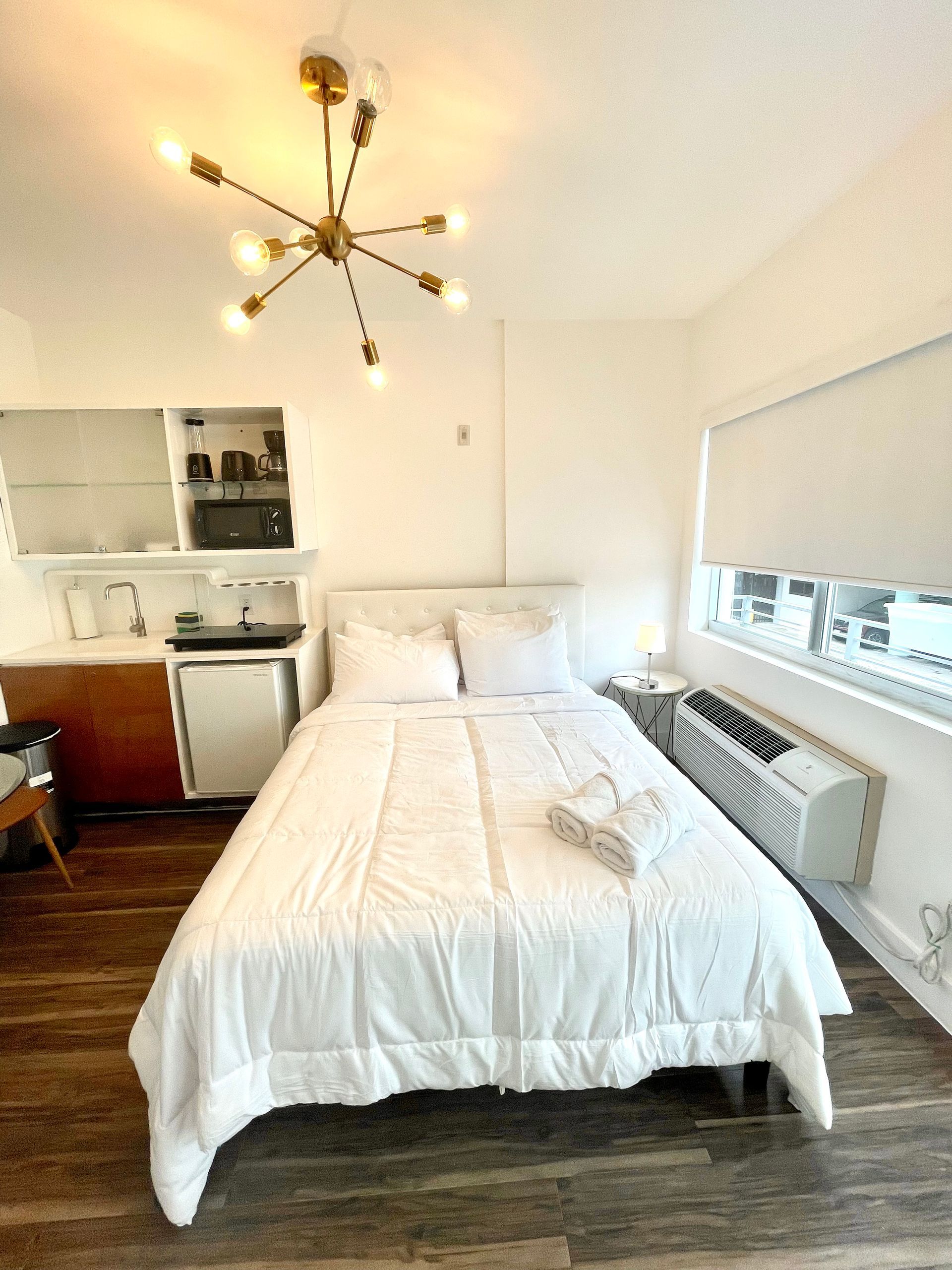 A small, bright studio apartment with a bed, kitchenette, and window with a partially lowered blind.