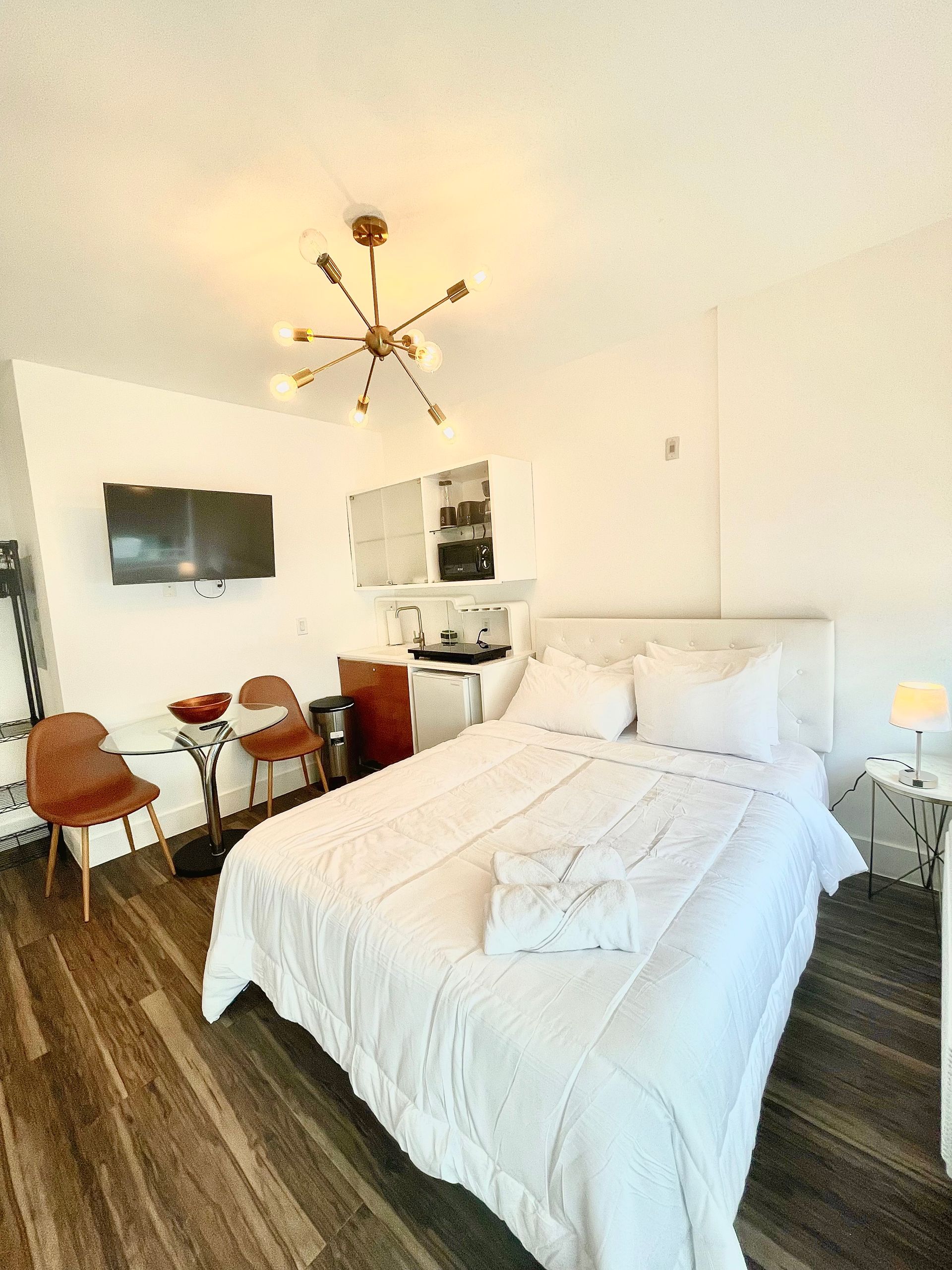 Cozy studio apartment with white bed, TV, kitchenette, chairs, and decorative chandelier.