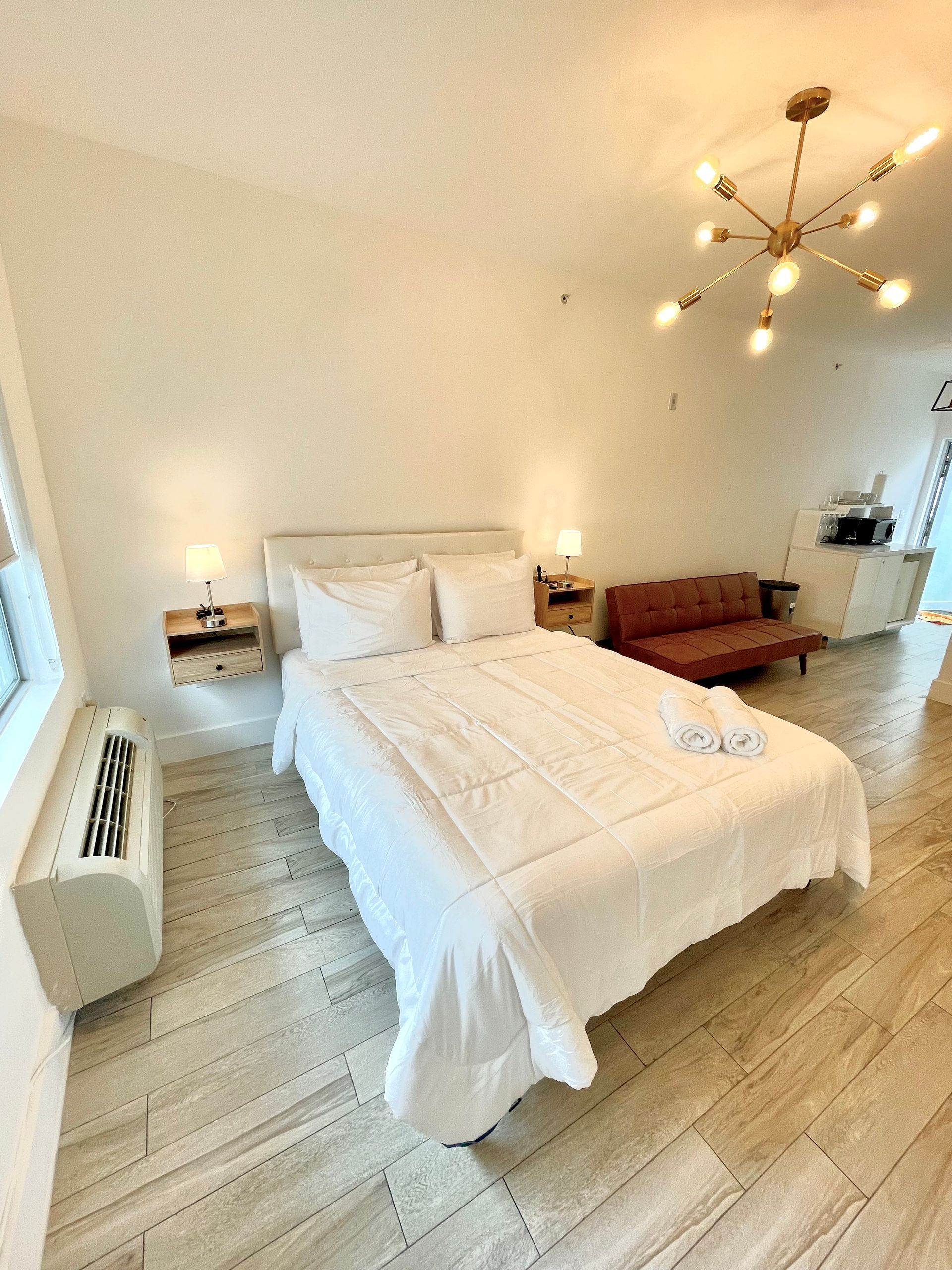 Bright bedroom with bed, couch, and modern light fixture. White walls, wooden floor, and air conditioning unit.